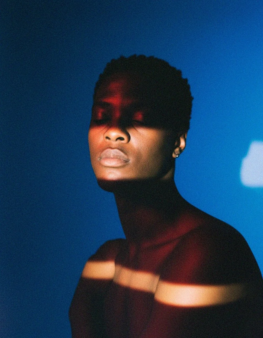 Portrait of a woman with short hair and earrings, with colored light and shadows cast on her face and shoulders against a blue background.