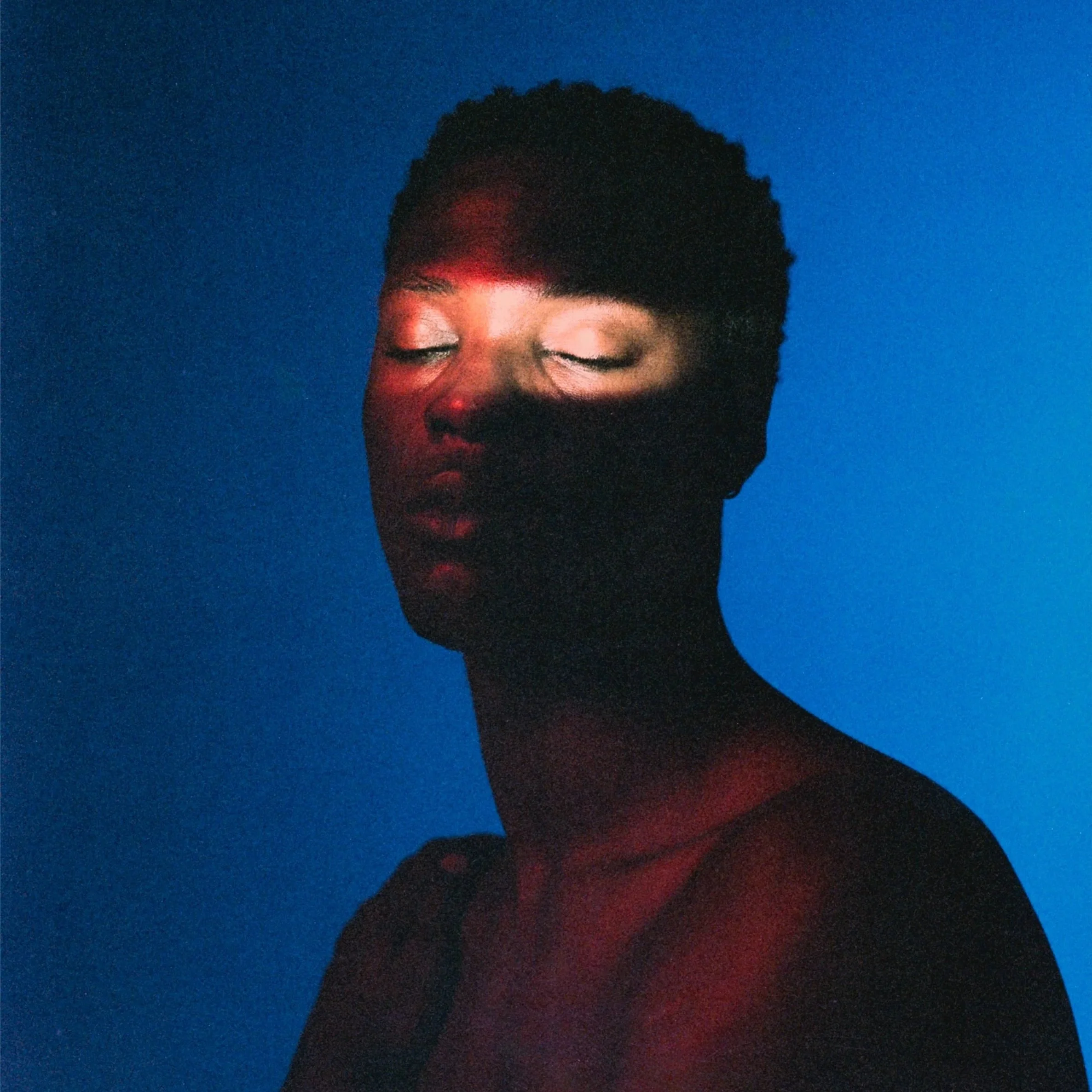 Close-up of a person with closed eyes and dark hair against a blue background, illuminated by soft red light.