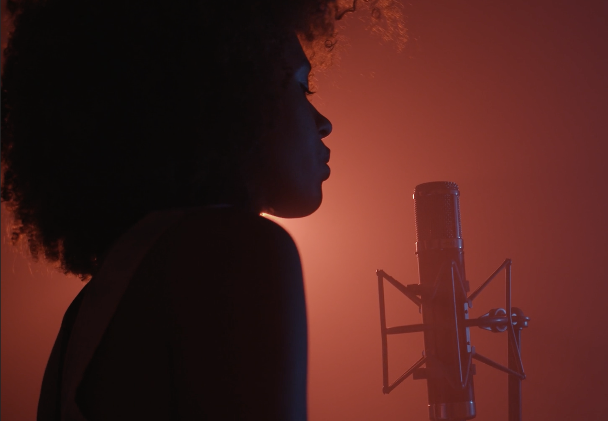 Silhouette of a person singing into a microphone against a red-orange background with broadcasting equipment.