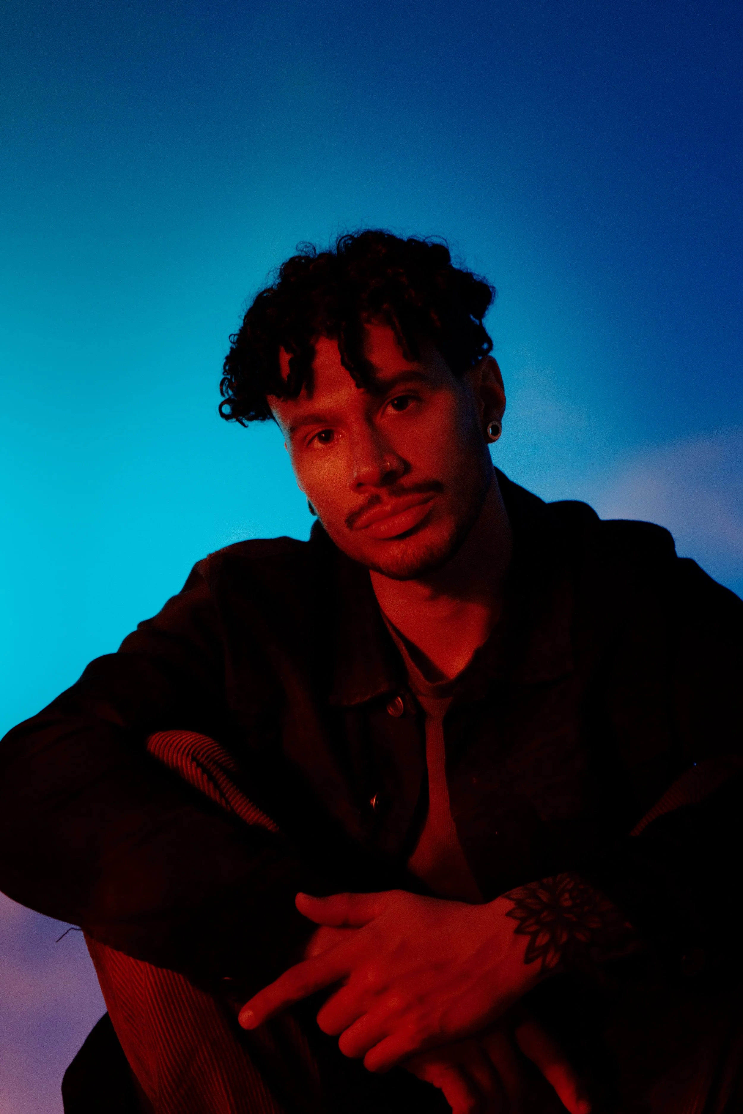 A young man with curly dark hair, earrings, and a nose ring, sitting with his hands folded in front of him, illuminated by red and blue lighting, with the sky in the background.
