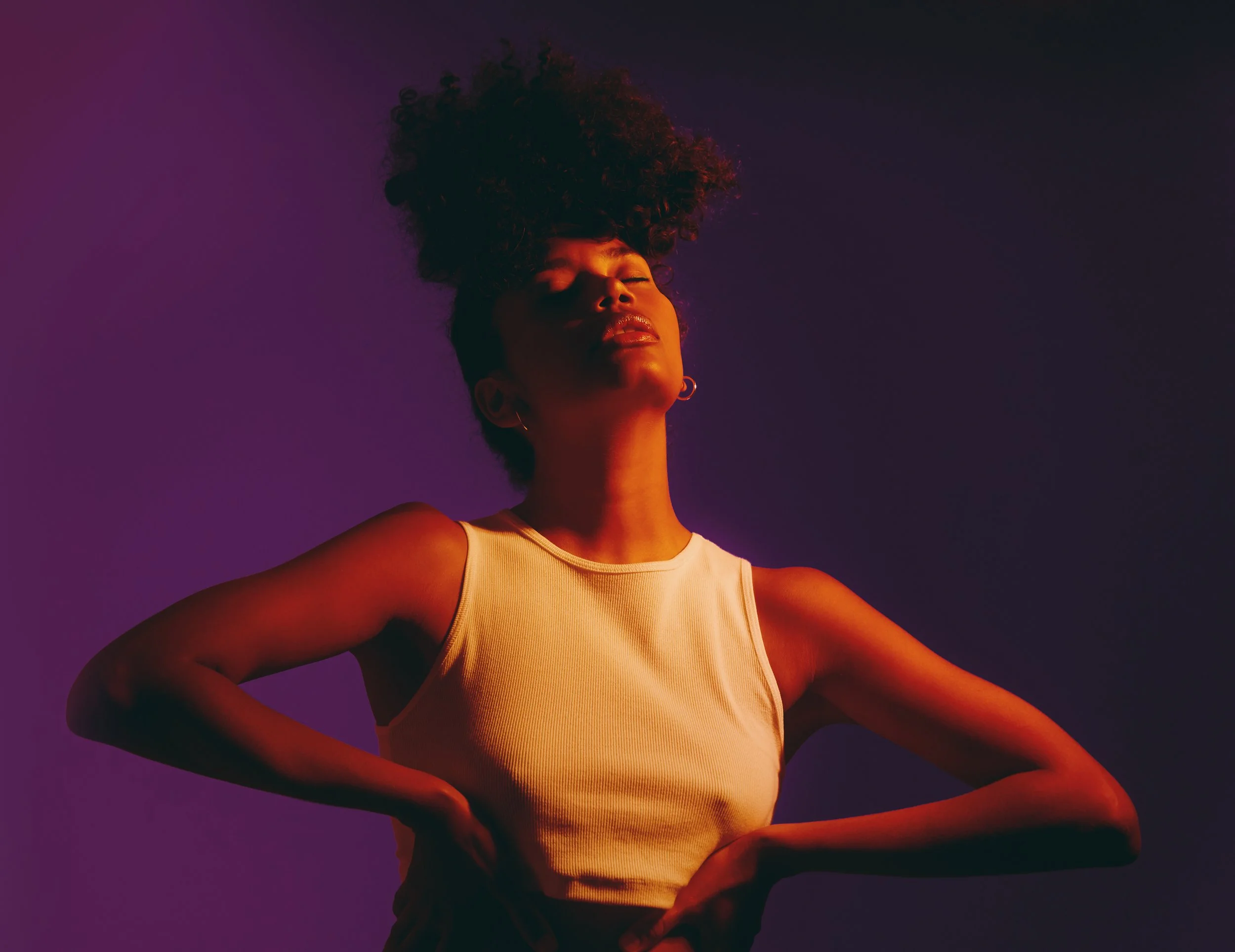 A woman with curly hair styled up, wearing a beige tank top, with her eyes closed and arms akimbo, was illuminated by colorful purple and orange lighting against a dark background.