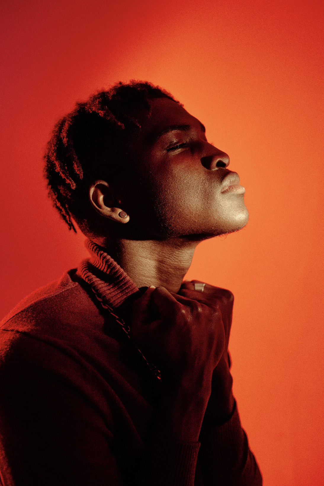 Portrait of a person with short twisted hair, wearing a dark jacket, with eyes closed and holding the collar with one hand, against a red background.