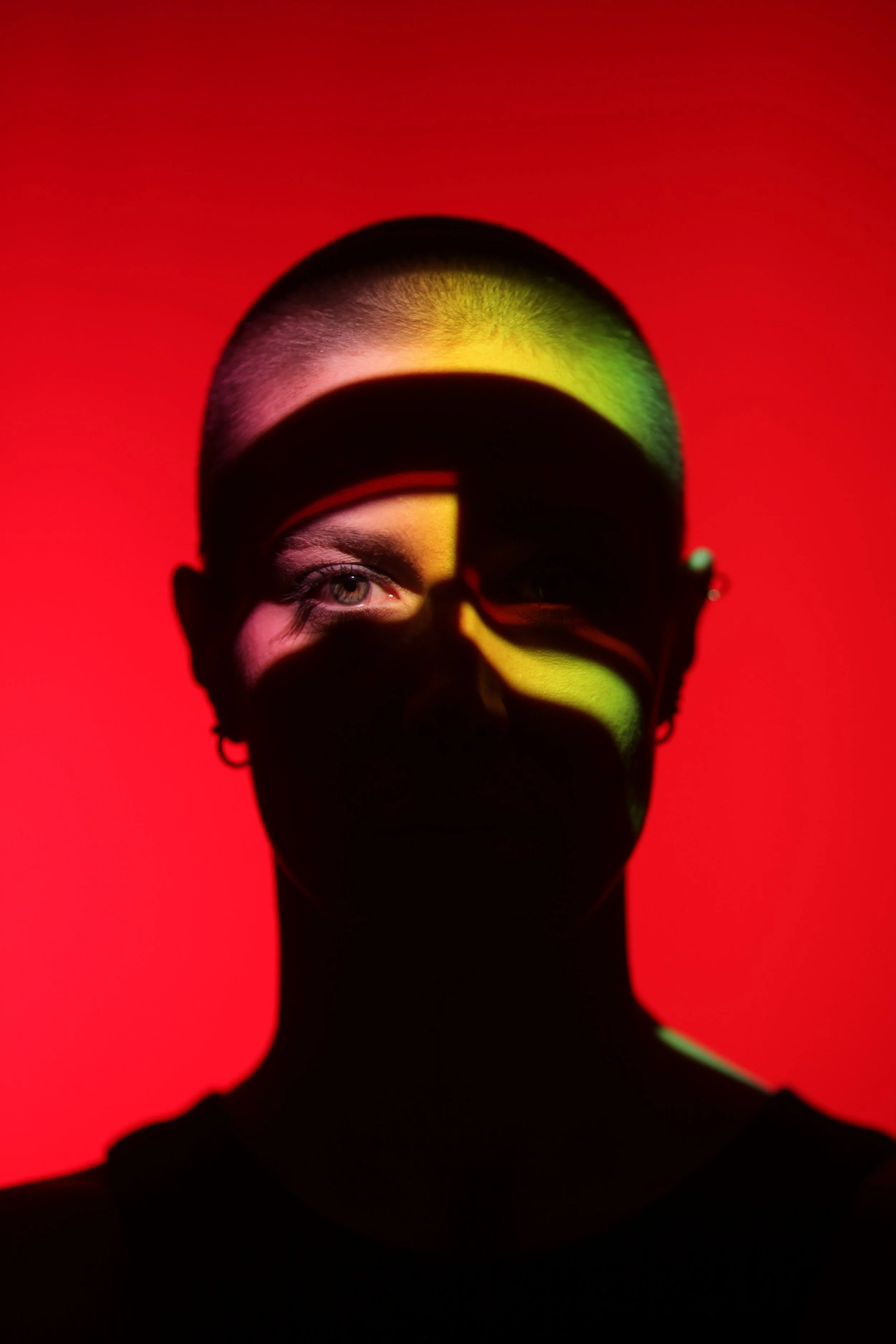 A person wearing glasses and a mask, with one eye visible, illuminated by colorful lighting against a red background.