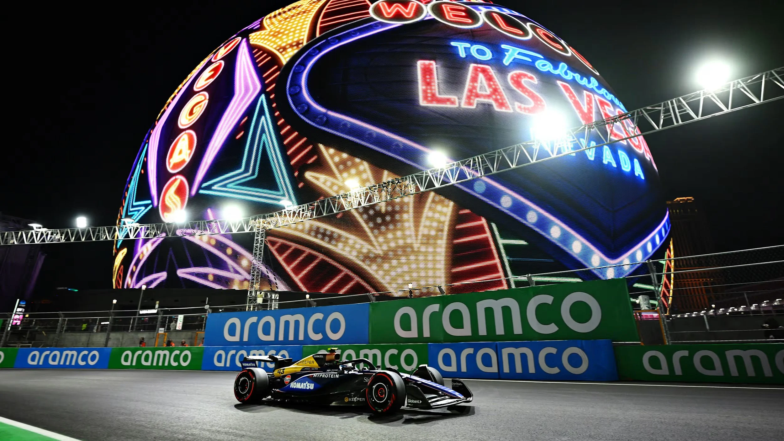 Nighttime view of a Formula 1 race car with sponsor logos on a race track, with a large neon-lit globe sign in the background displaying Las Vegas and the words 'To Fabulous Las Vegas'.