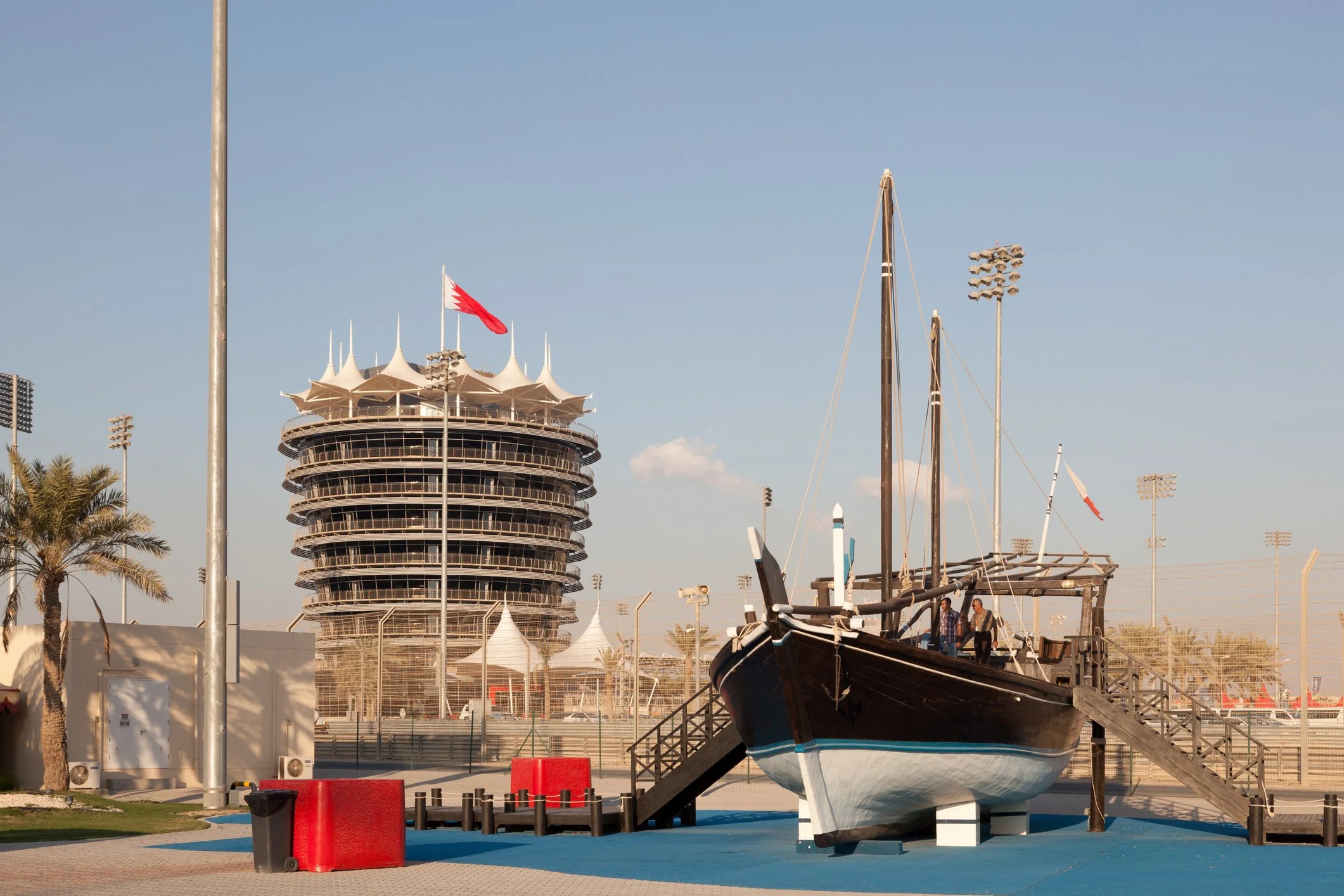 A sailboat on dry land next to a dock with masts and a building with a futuristic design featuring multiple pointed roofs and a red and white flag, near palm trees and stadium lights.