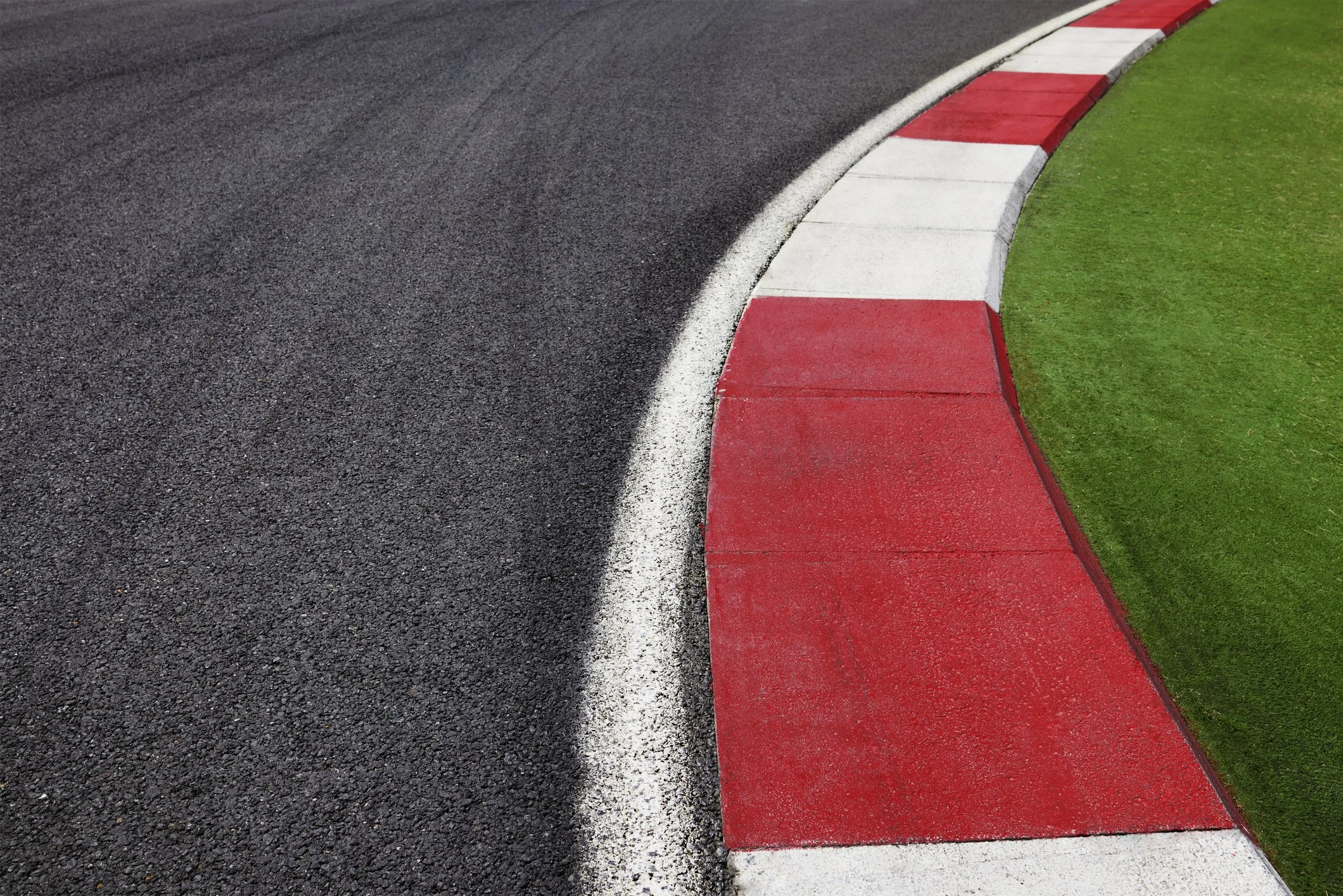 A close-up view of a race track curb with red and white painted sections on the edge of the asphalt racing surface, next to a patch of green grass.