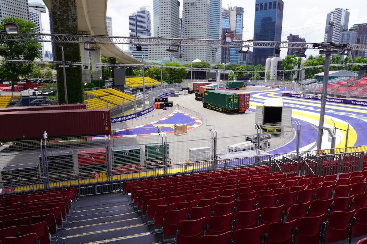 View of a Formula 1 racetrack set up in an urban city with tall buildings, empty red seating, and trucks on the track preparing for a race.