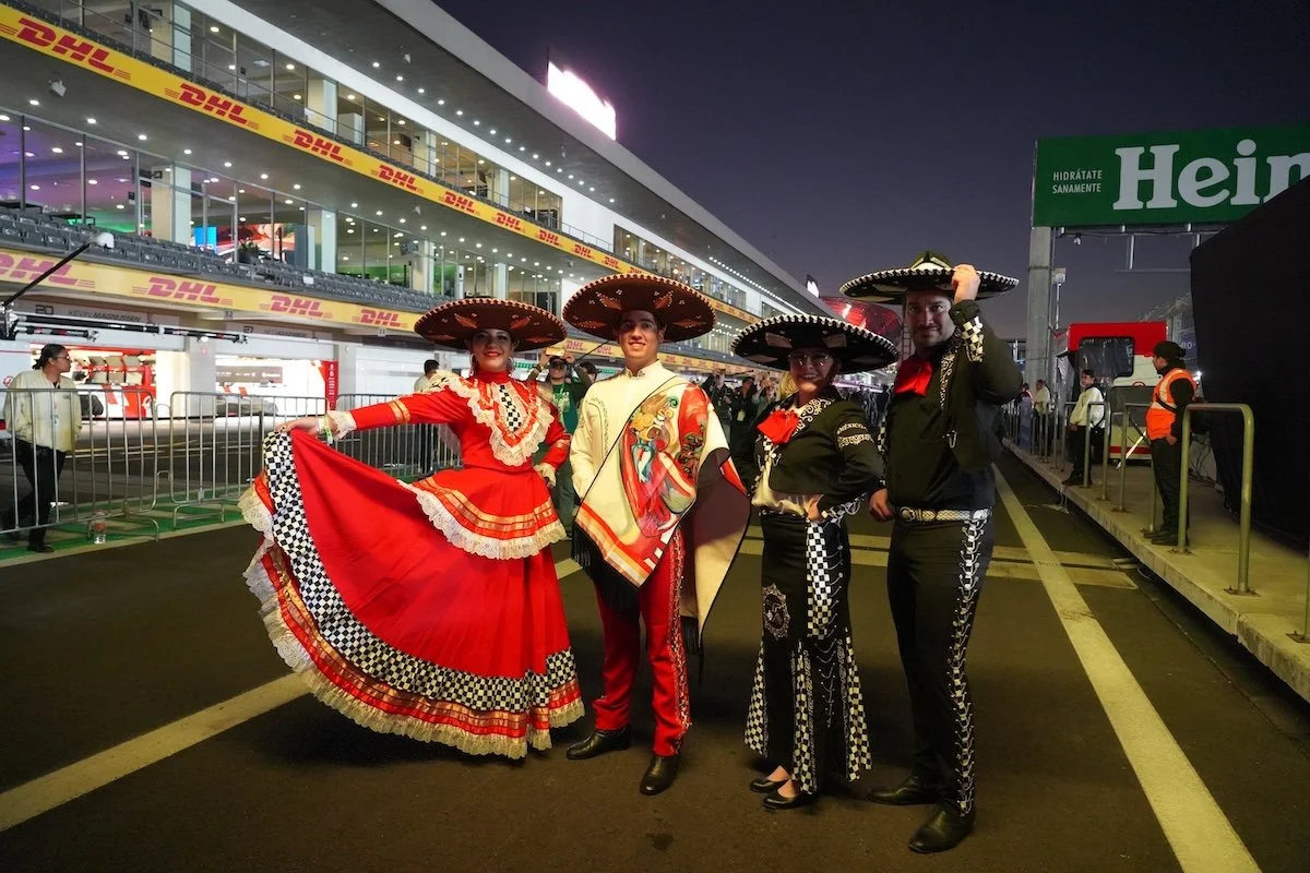 Group of four people dressed in traditional Mexican Mariachi and folkloric costumes, standing in a parking area at night with a building and DHL banners in the background.