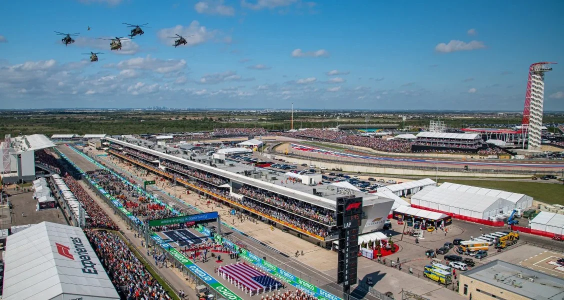 Race track at a motorsports event with grandstands crowded with spectators, a pit lane, and a helicopter flying overhead.