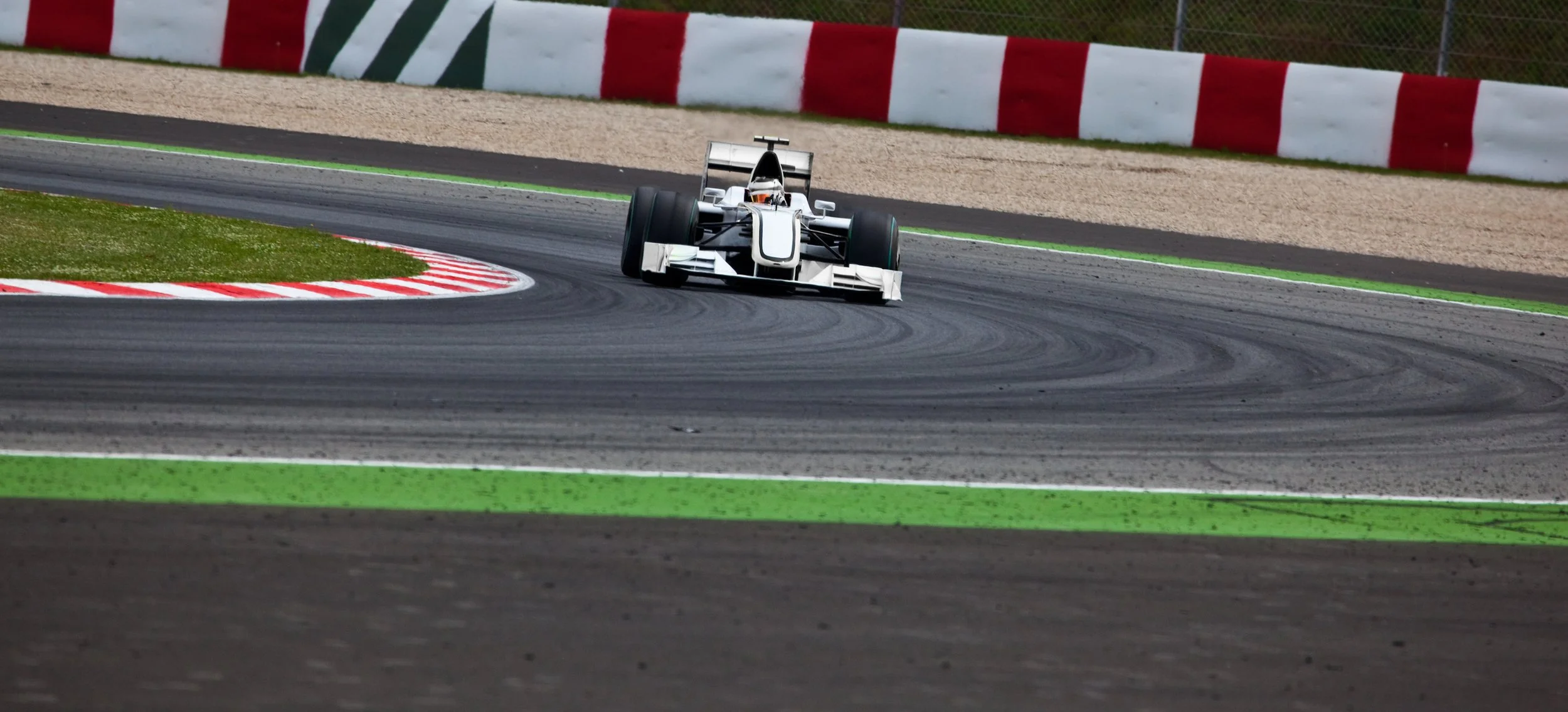 Race car on a racetrack, taking a curve, with red and white barriers in the background and green painted lines on the track.
