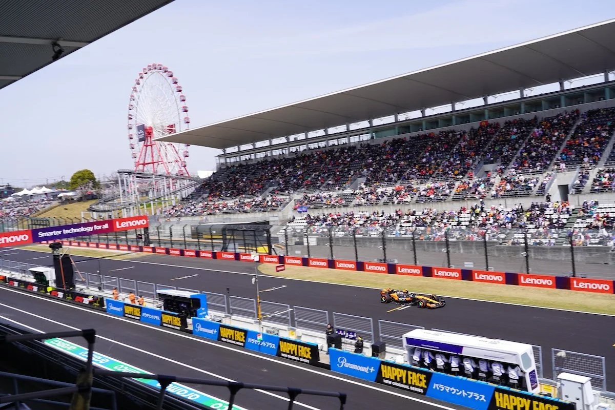 Race track with a car speeding past, grandstands full of spectators, a ferris wheel in the background, and advertising banners lining the track.