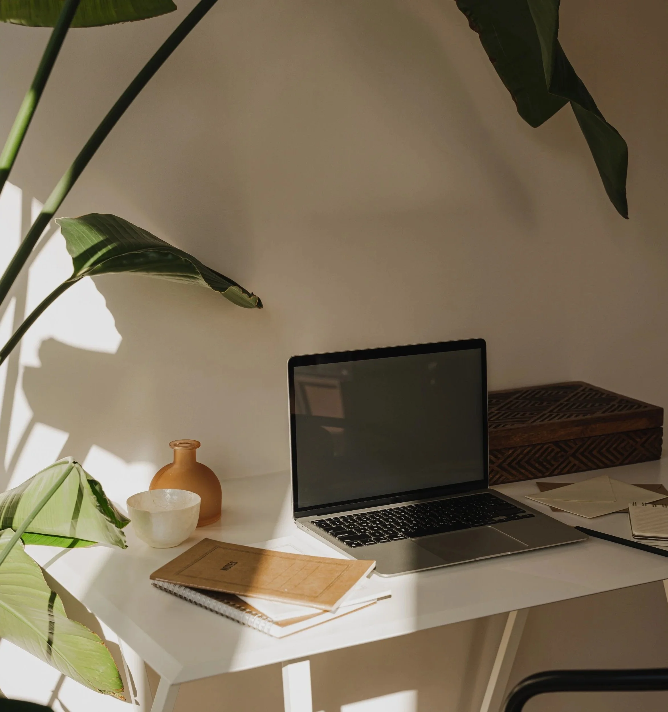 A white desk with a silver laptop, a light brown notebook, a darker brown spiral notebook, a white ceramic bowl, a small terracotta vase, some papers, and a green potted plant on the left side of the desk.