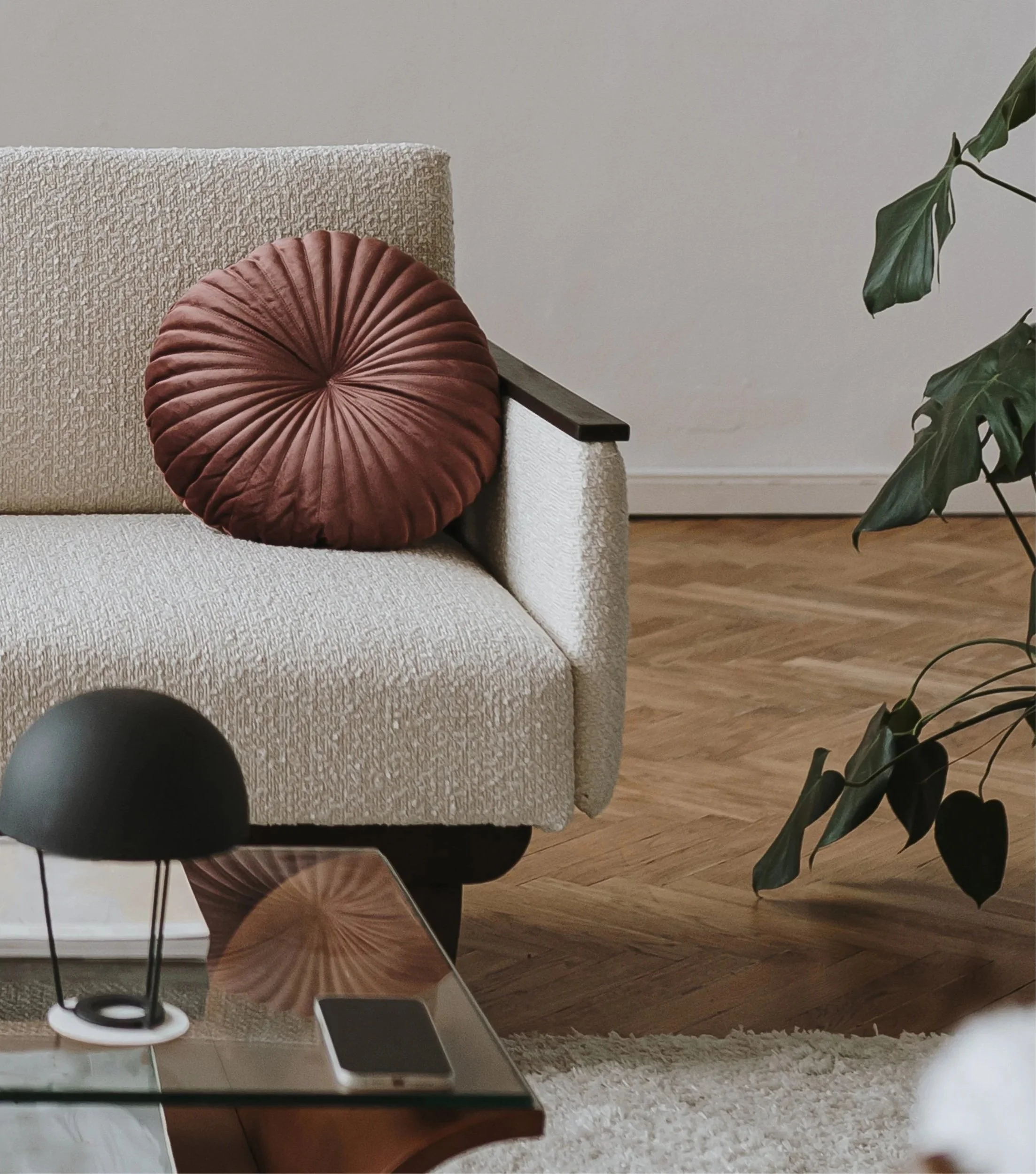 Living room with a beige textured sofa, a round rust-colored velvet cushion, a potted plant with green leaves on the right, a wooden floor, a white wall, a table with a smartphone, and a floor lamp.