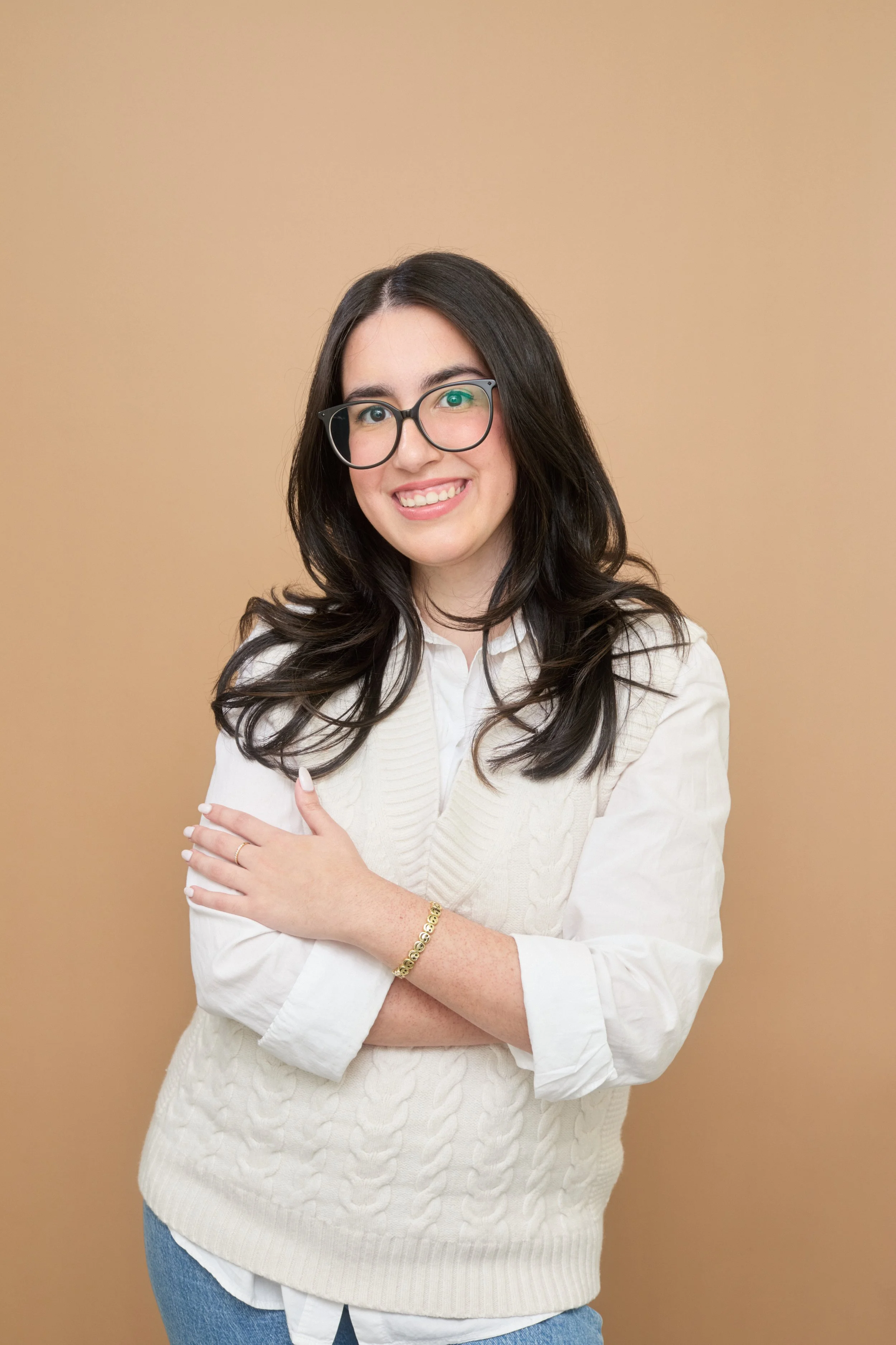 A therapist with long dark hair, glasses, and light skin, smiling and crossing her arms, wearing a white sweater over a collared shirt and blue jeans, standing against a beige background.