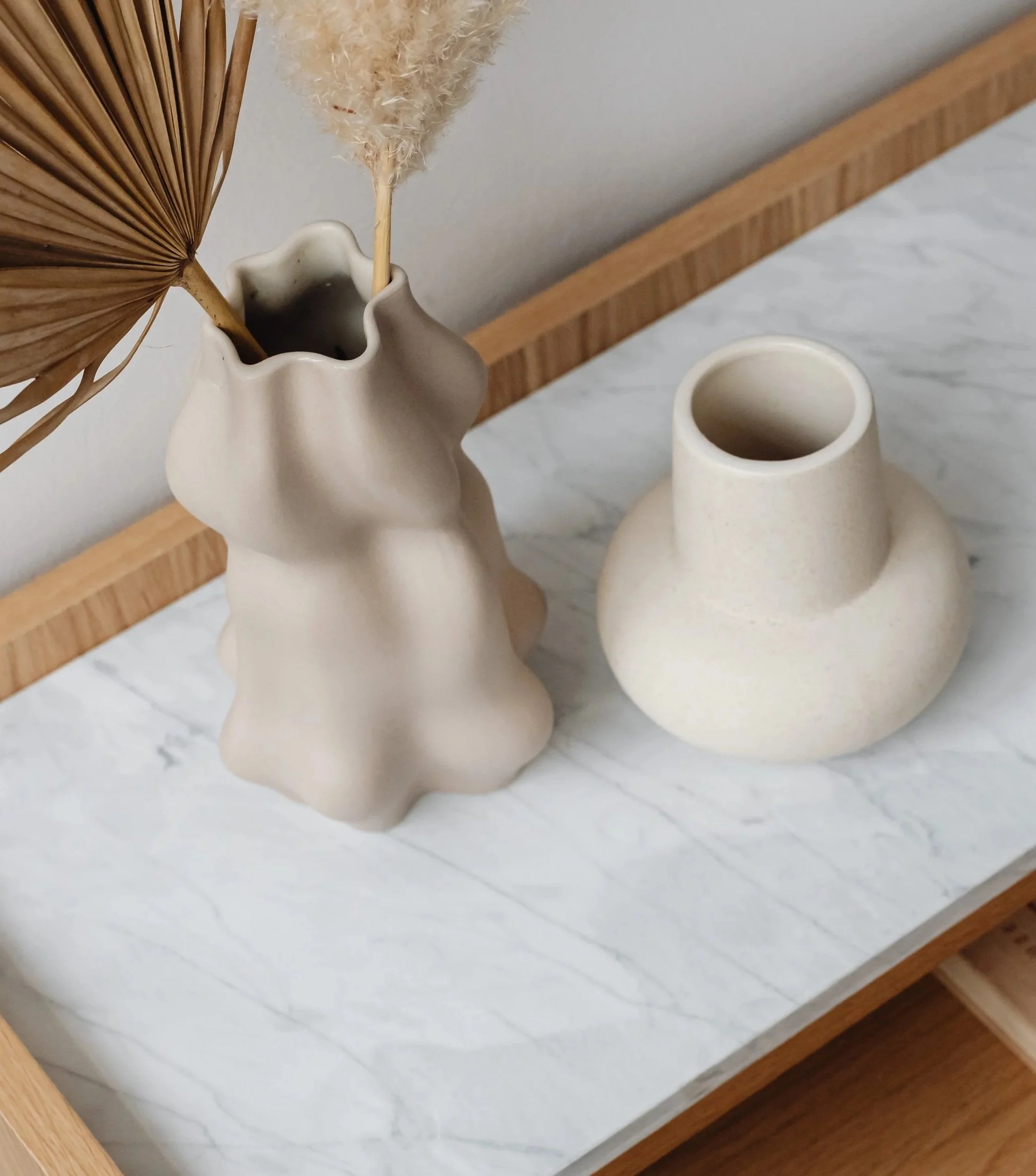 Close-up photo of two beige ceramic vases with narrow openings on a light-colored marble surface, with one vase holding dried pampas grass and a what appears to be a dried palm leaf, against a background of a wooden frame and plain off-white wall.