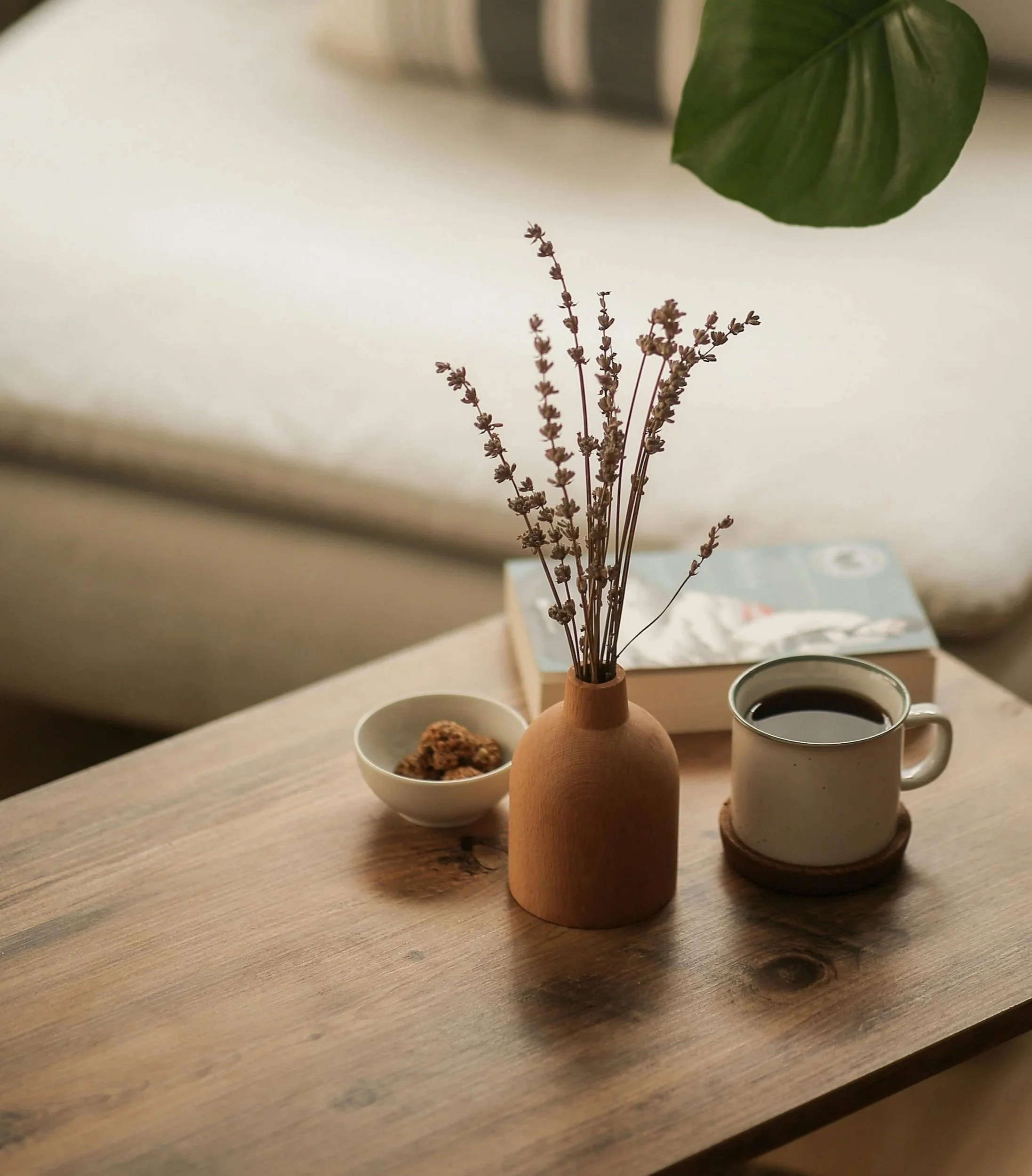 A small wooden vase with dried lavender and a large green leaf above it, accompanied by a white bowl with baked goods and a white mug of coffee on a wooden table.