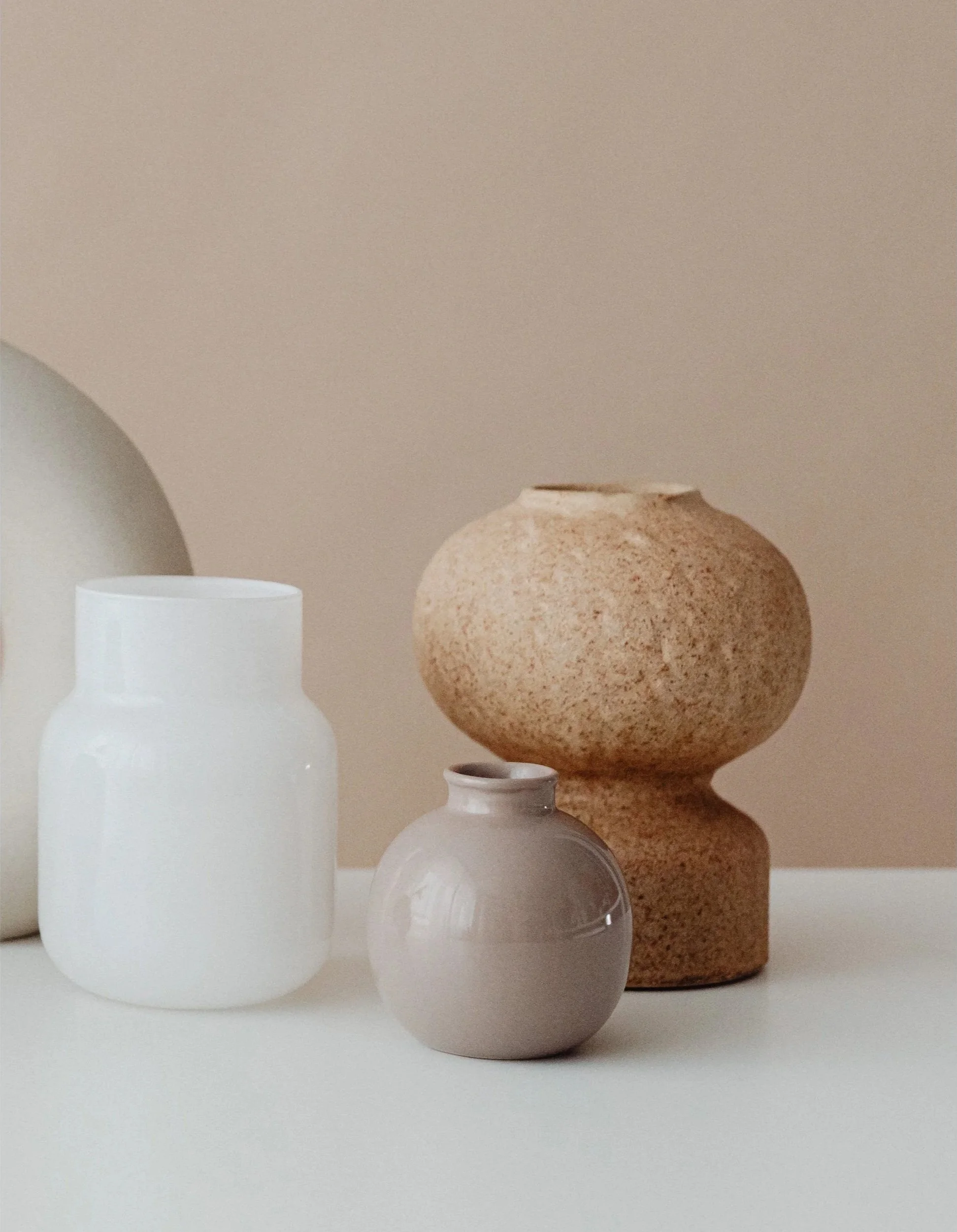 Four vases of different sizes and colors arranged on a white surface against a beige wall. The vases are minimalist and vary in shape, with one being tall and white, one small and terracotta-colored, and two others with rounded shapes.