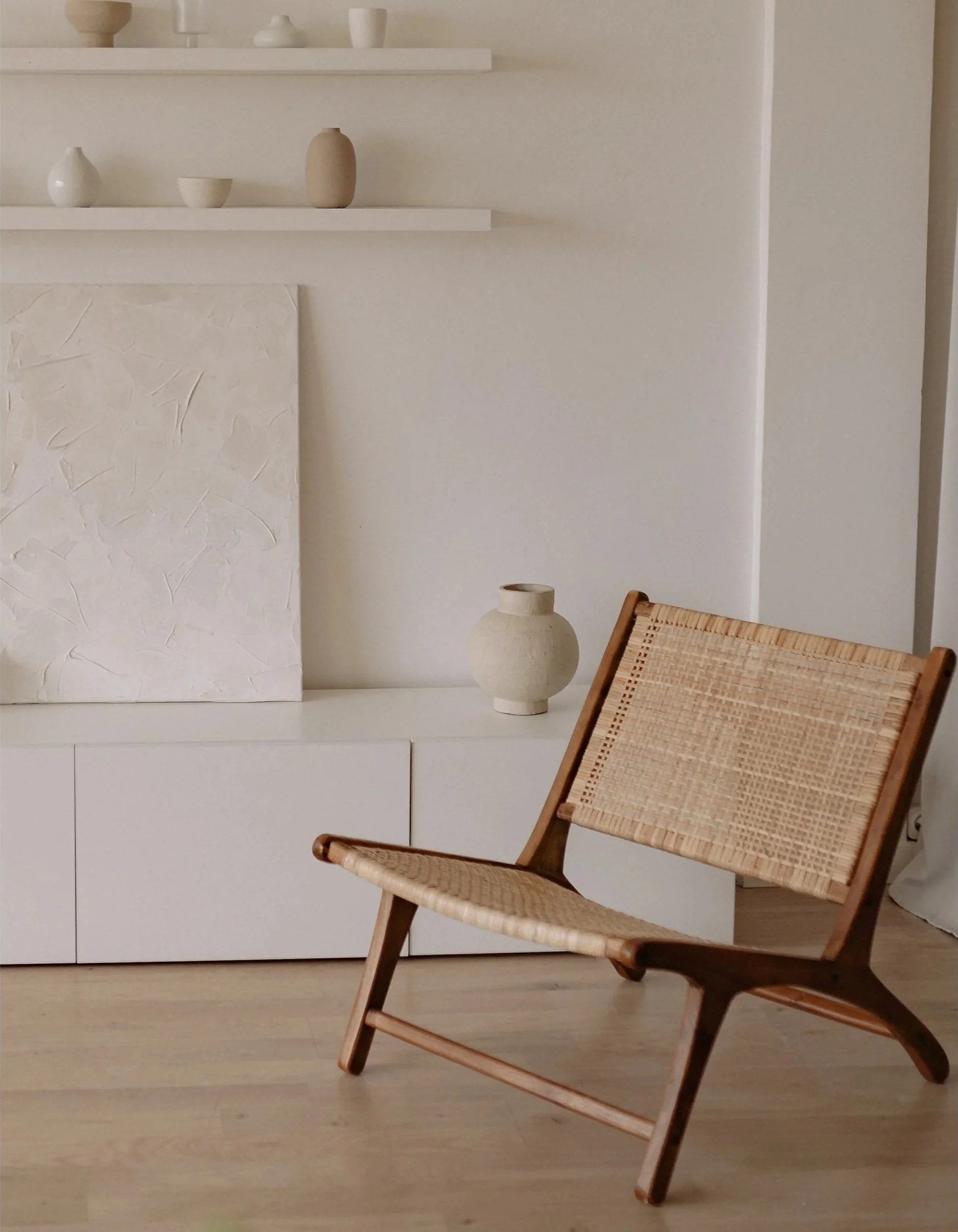 Interior scene with a wooden chair with woven seat and back, a white cabinet, decorative vases on a shelf, and a textured white panel on the wall.