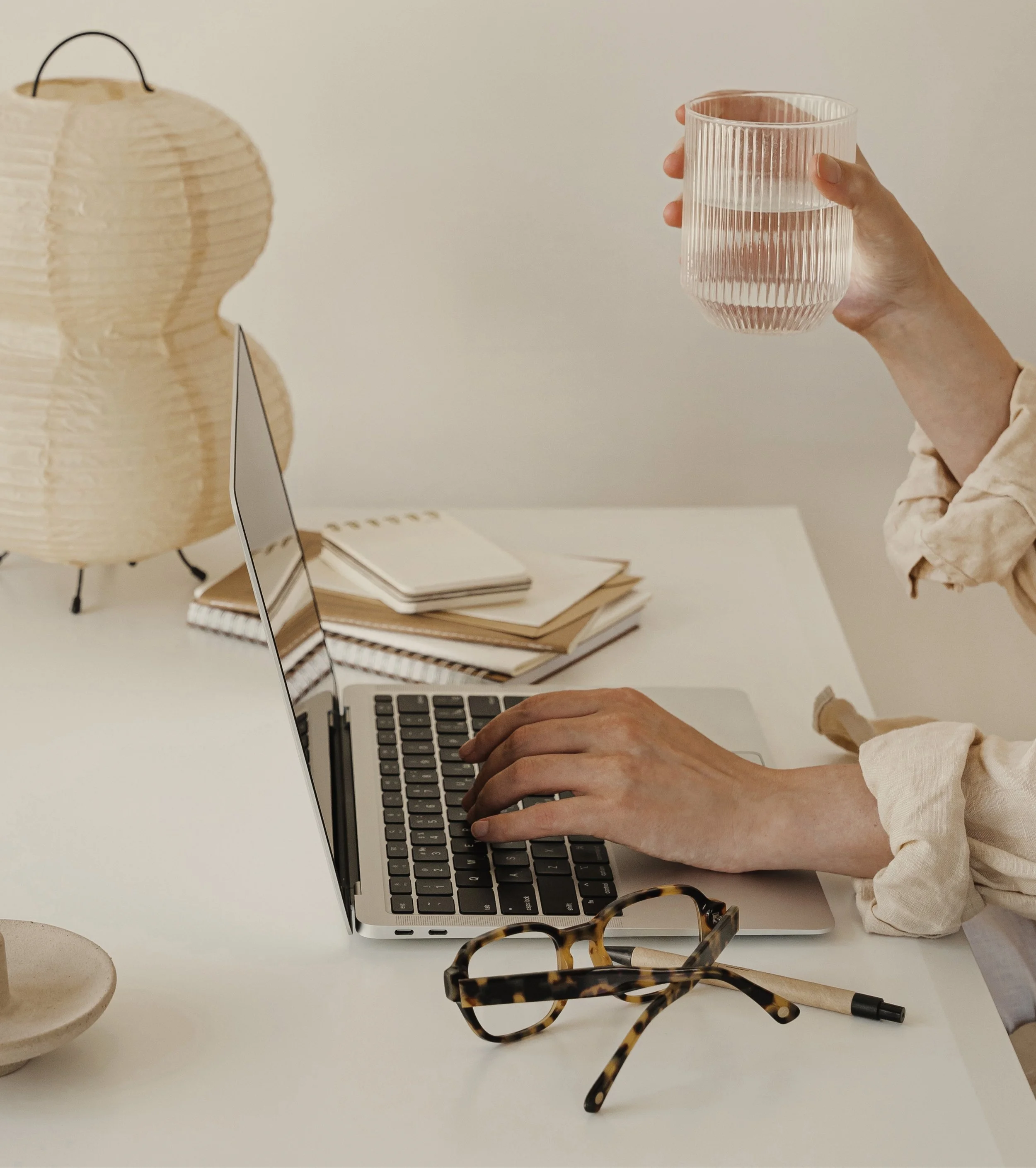 Person working on a laptop at a white desk, holding a glass of water, with notebooks, a paper lantern, glasses, a pen, and a bowl nearby.