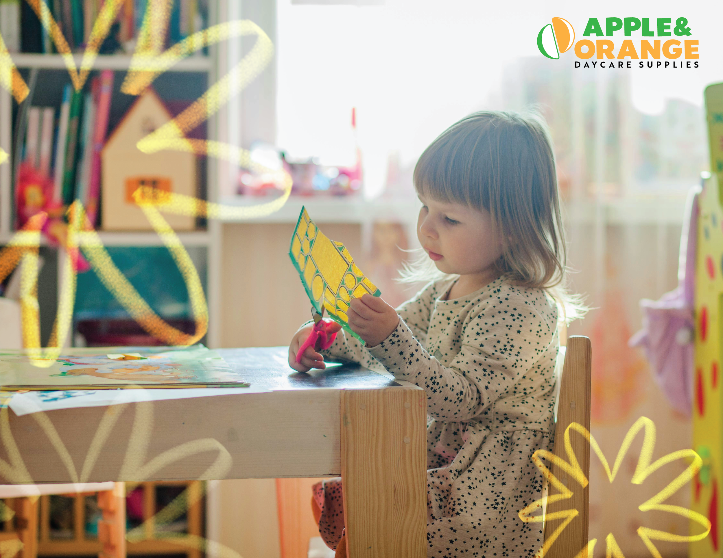 A young girl with light to medium brown hair, wearing a beige shirt with black star patterns, sitting at a wooden table, cutting a piece of yellow patterned paper with pink scissors in a room with natural light and colored paper decorations.