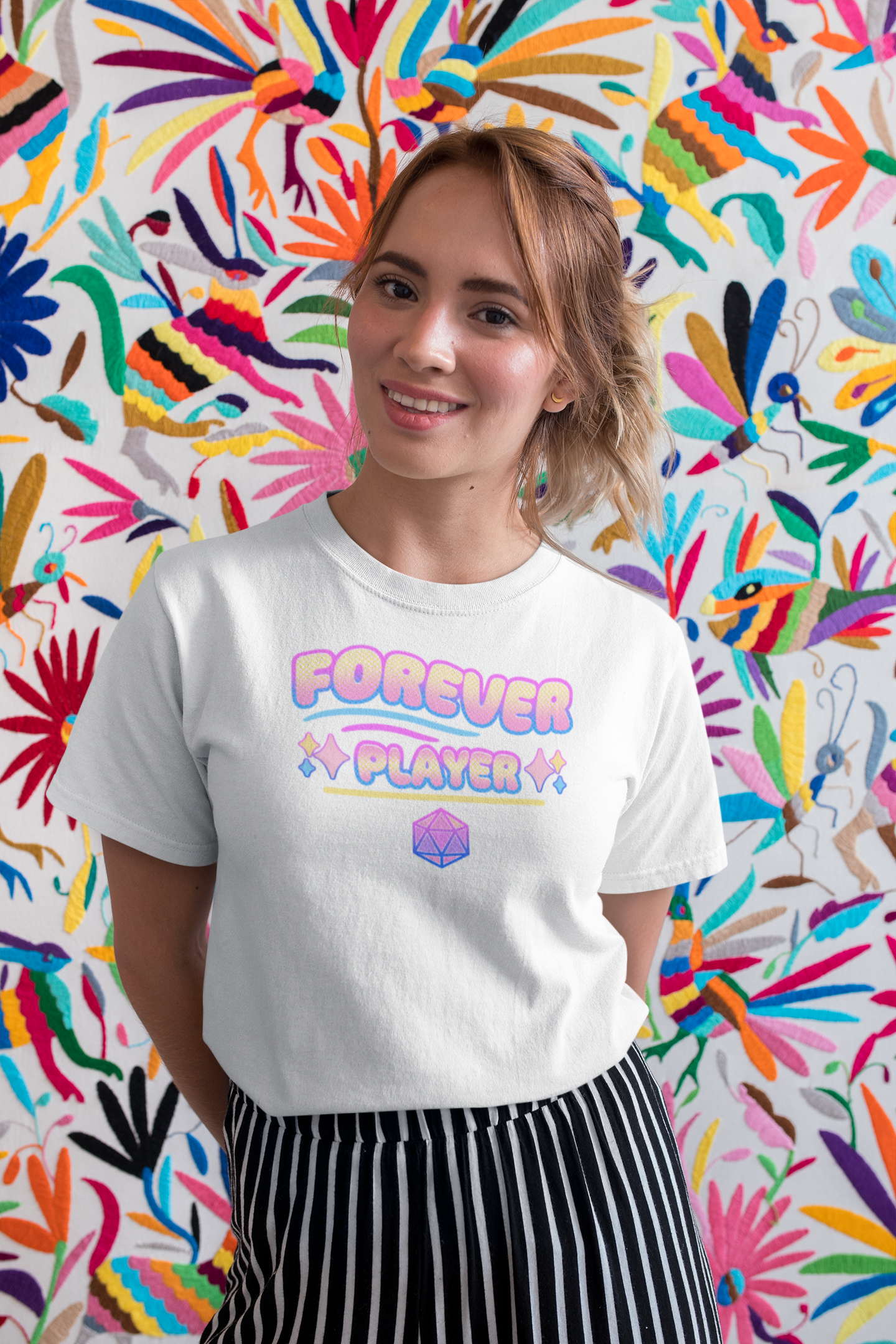A young woman with short, wavy blonde hair and light skin is standing in front of a colorful decorative backdrop. She is smiling and wearing a white T-shirt with pastel-colored text that reads "FOREVER PLAYER" with a geometric design below. She has small earrings and is dressed in black and white striped pants.