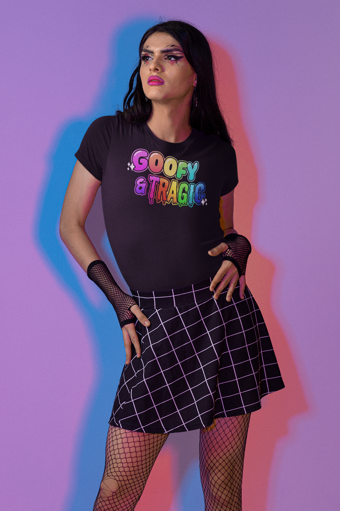 Person wearing a black T-shirt with colorful text that says 'Goofy & Tragic,' black fishnet stockings, and a plaid skirt, standing against a multicolored background.