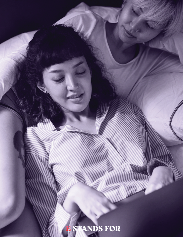 Two women lying in bed; one with dark curly hair, the other with short light hair, looking at a tablet. Black and white photo.