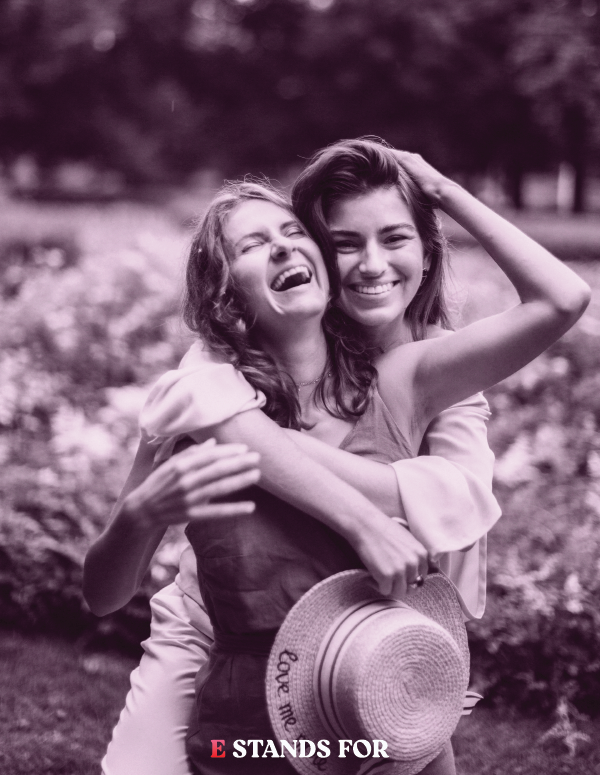 Two young women are hugging and smiling outdoors in a park, with one holding a straw hat that has the word 'Love' embroidered on the brim.