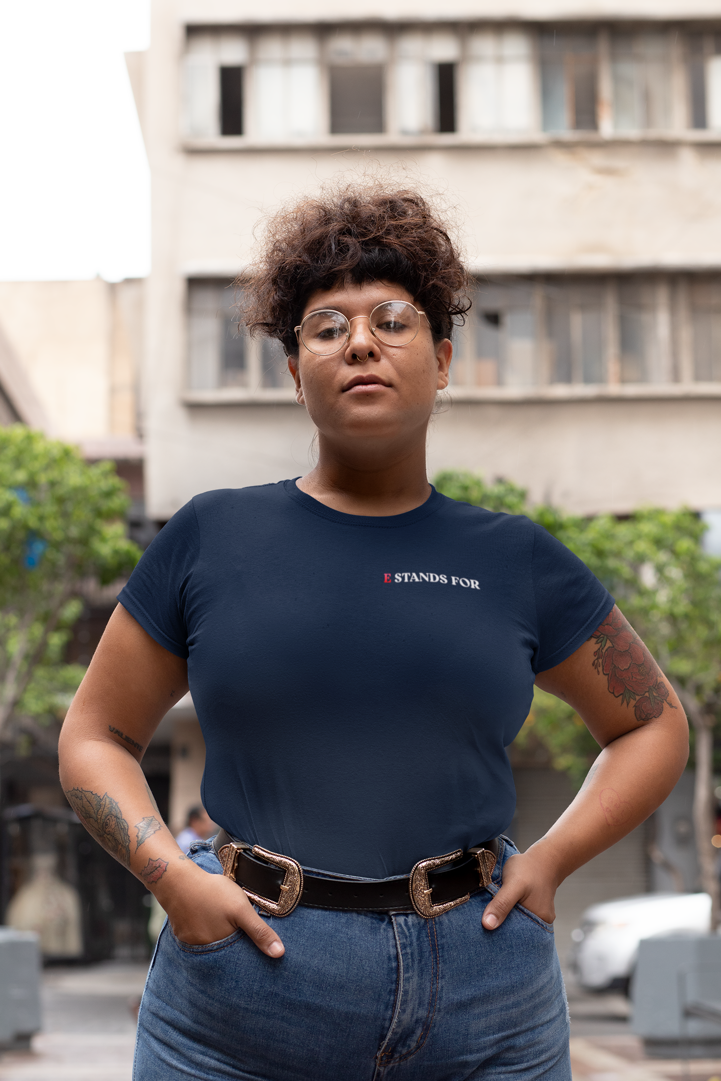 A person with short curly hair, glasses, tattoos, and wearing a dark blue T-shirt and jeans, standing outdoors with a building and trees in the background.