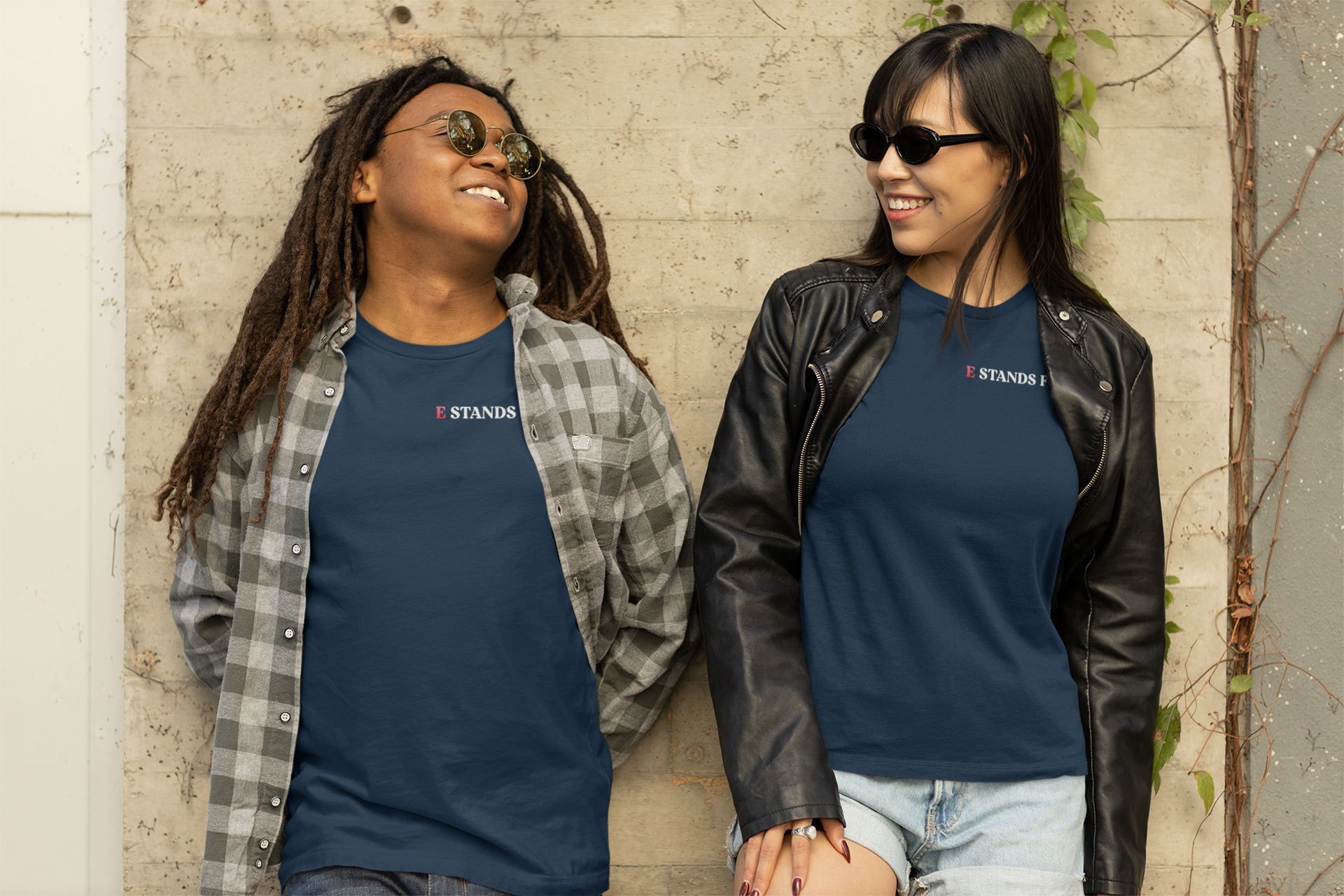 Two women standing close together against a beige wall, smiling at each other. They are wearing sunglasses and casual clothing, one in a plaid shirt over a T-shirt and the other in a leather jacket over a T-shirt.