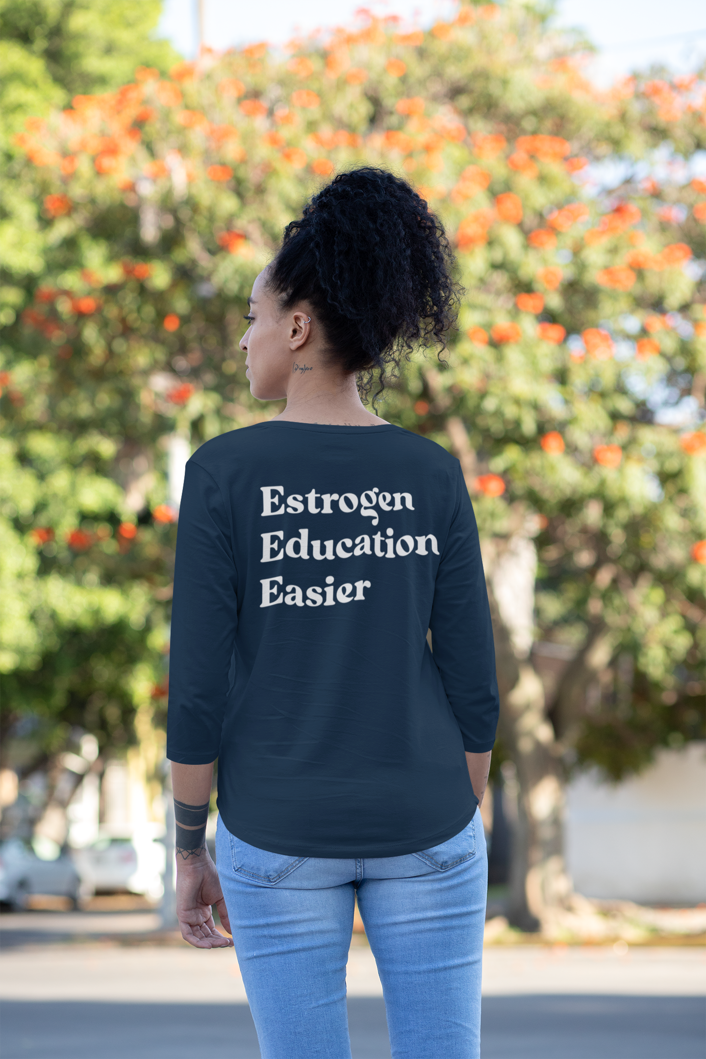 A woman with curly black hair in a high puff, wearing a navy blue long sleeve shirt with white text that reads 'Estrogen Education Easier' on the back. She is outdoors with a blurred background of green trees and orange flowers.