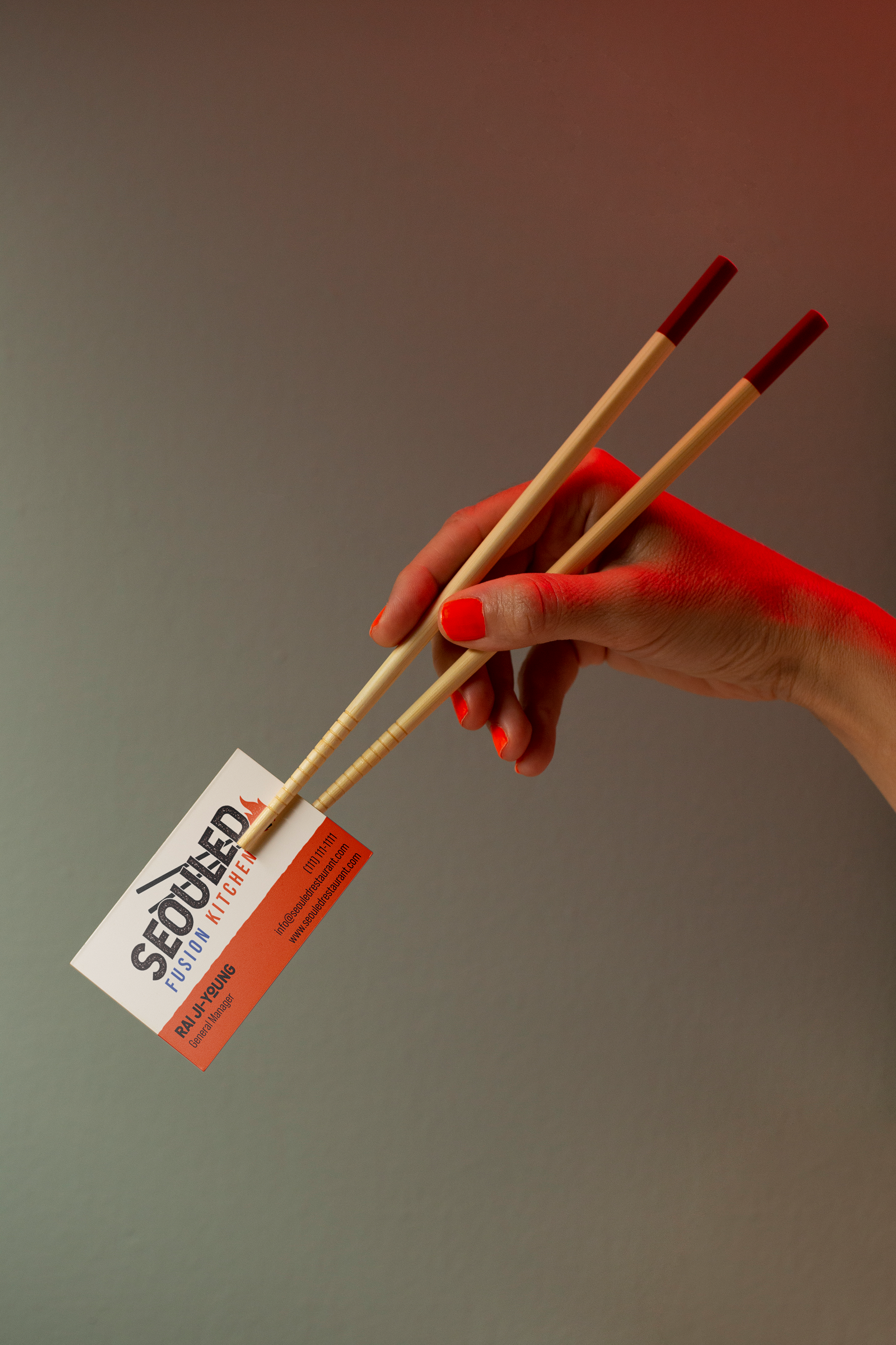 A person with red painted nails holding two beige chopsticks with red tips, and a business card that reads 'SEOULEDUCATION FUSION KITCHEN' in black and orange text.