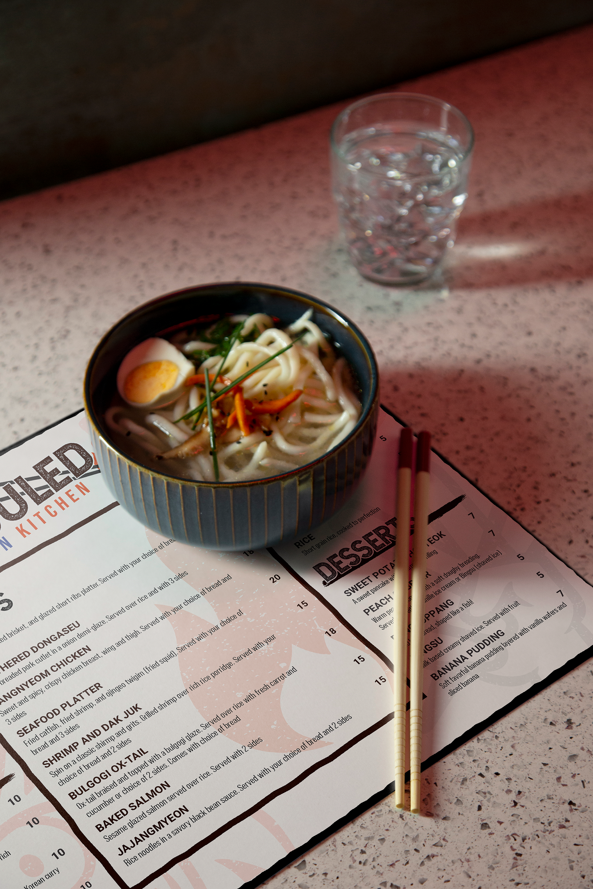 A bowl of noodle soup with sliced meat, a boiled egg, and green herbs on a menu with chopsticks on a pink speckled countertop.