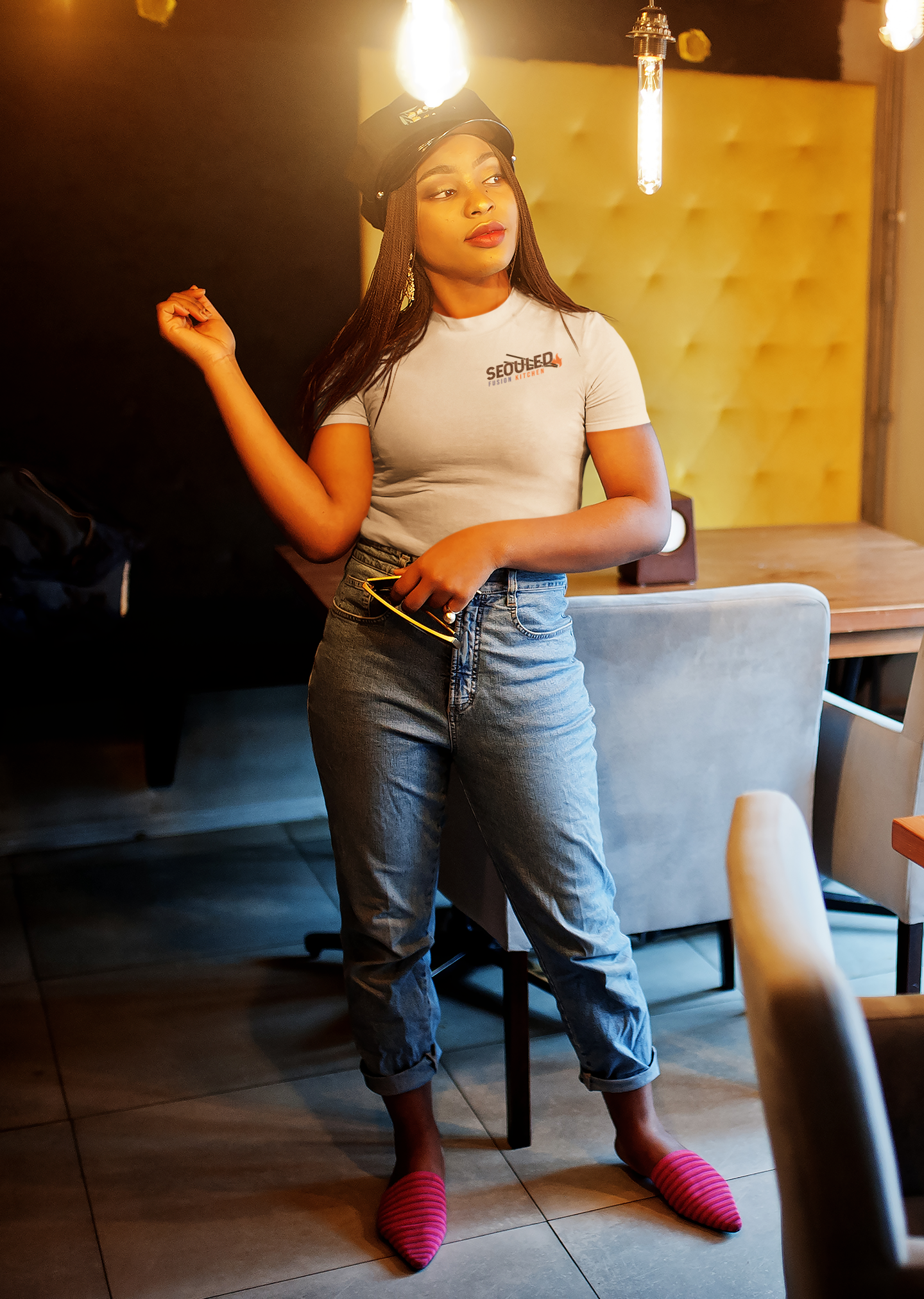 Young woman with long hair wearing a cap and a T-shirt with the text 'Seoul Ted Fusion Kitchen,' jeans, and pink striped slippers, standing in a restaurant with modern lighting and furniture.