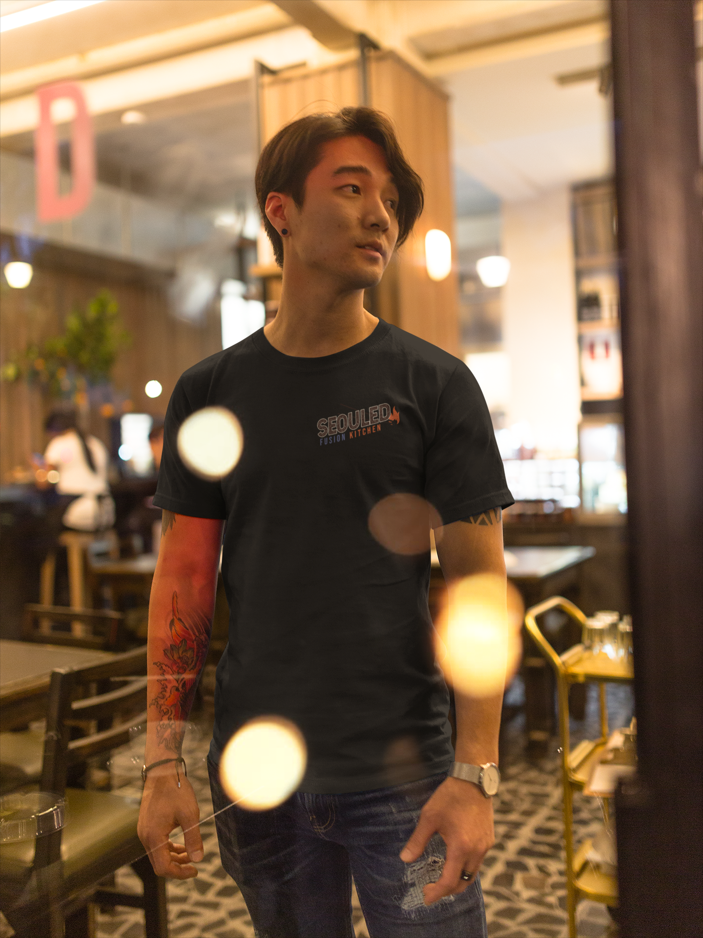 A young man with dark hair and tattoos on his arm, wearing a black T-shirt with 'SEOUL' printed on it, stands in a warmly lit restaurant or cafe with wood accents and patterned carpet. He has a watch on his wrist and a ring on his finger, and is looking slightly to his right.