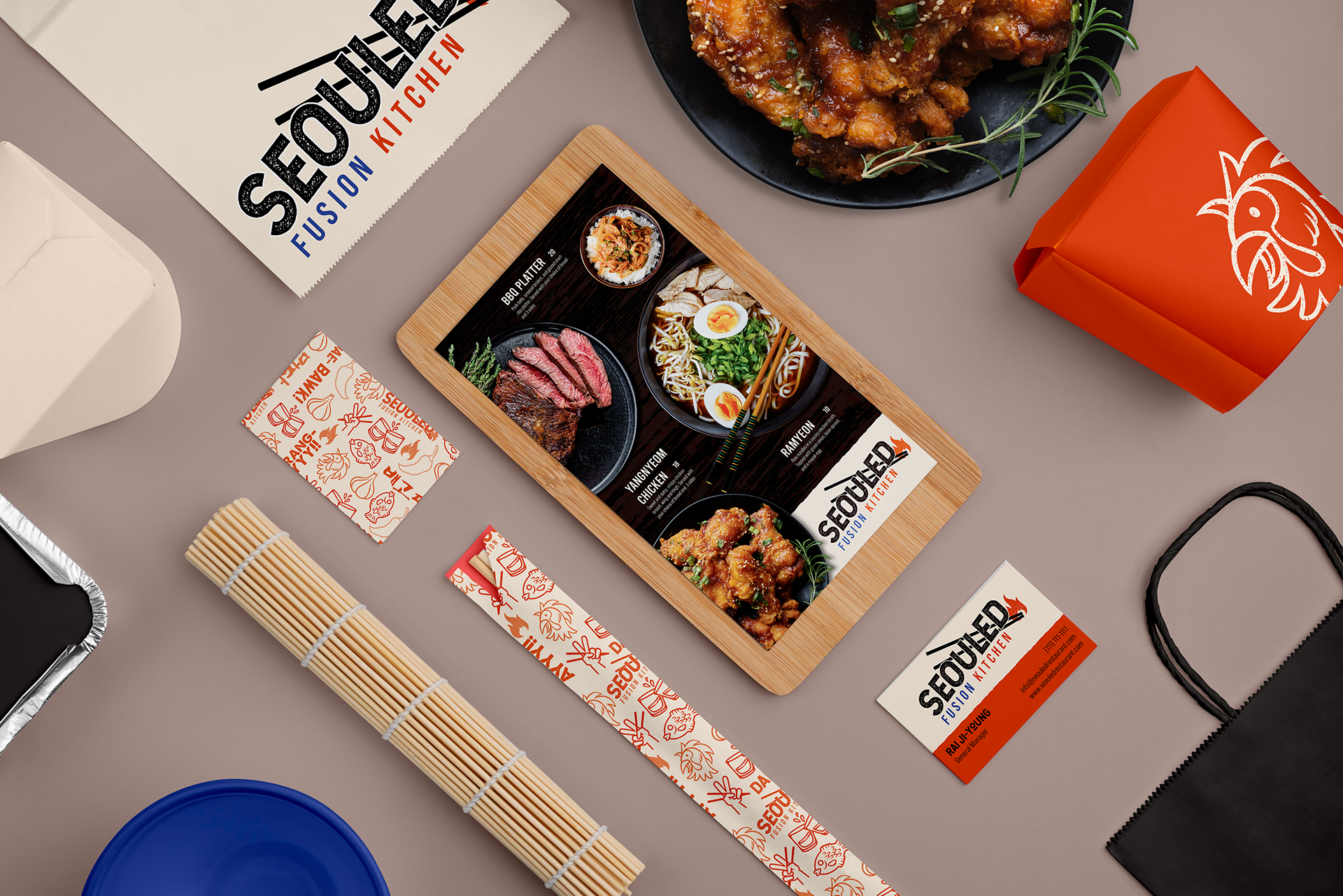 Top-down view of Asian cuisine takeout items including a menu, a box of fried chicken, a bowl of ramen, a business card, and chopsticks on a gray surface.
