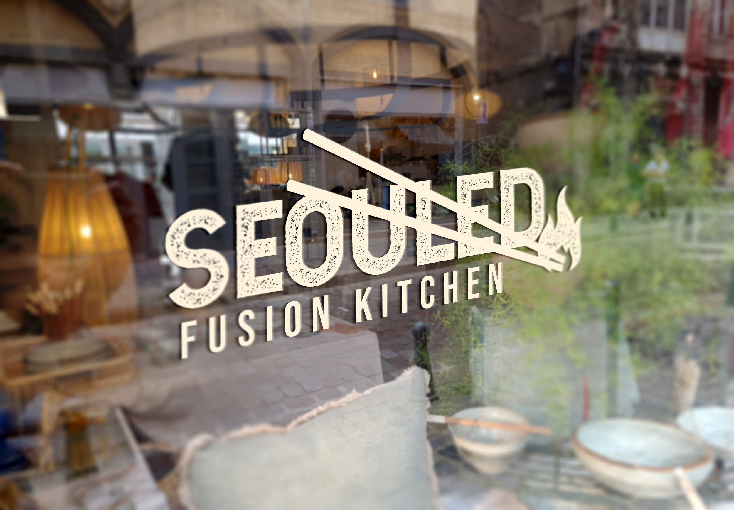 View through a window showing a sign for a fusion kitchen, with outdoor seating and greenery visible outside.