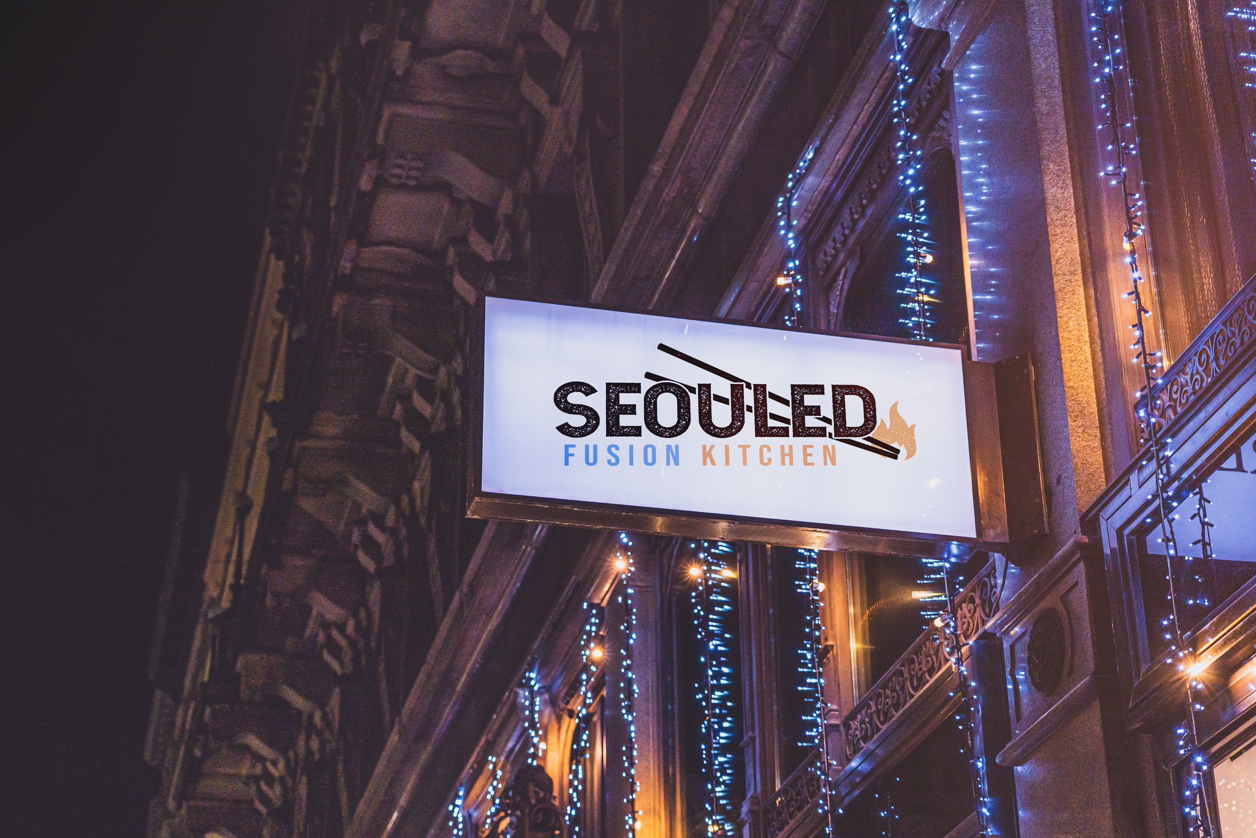 A illuminated sign on a building reads 'Seoul'ed Fusion Kitchen' with a silhouette of a fish and chopsticks. The building has ornate architectural details and blue string lights.