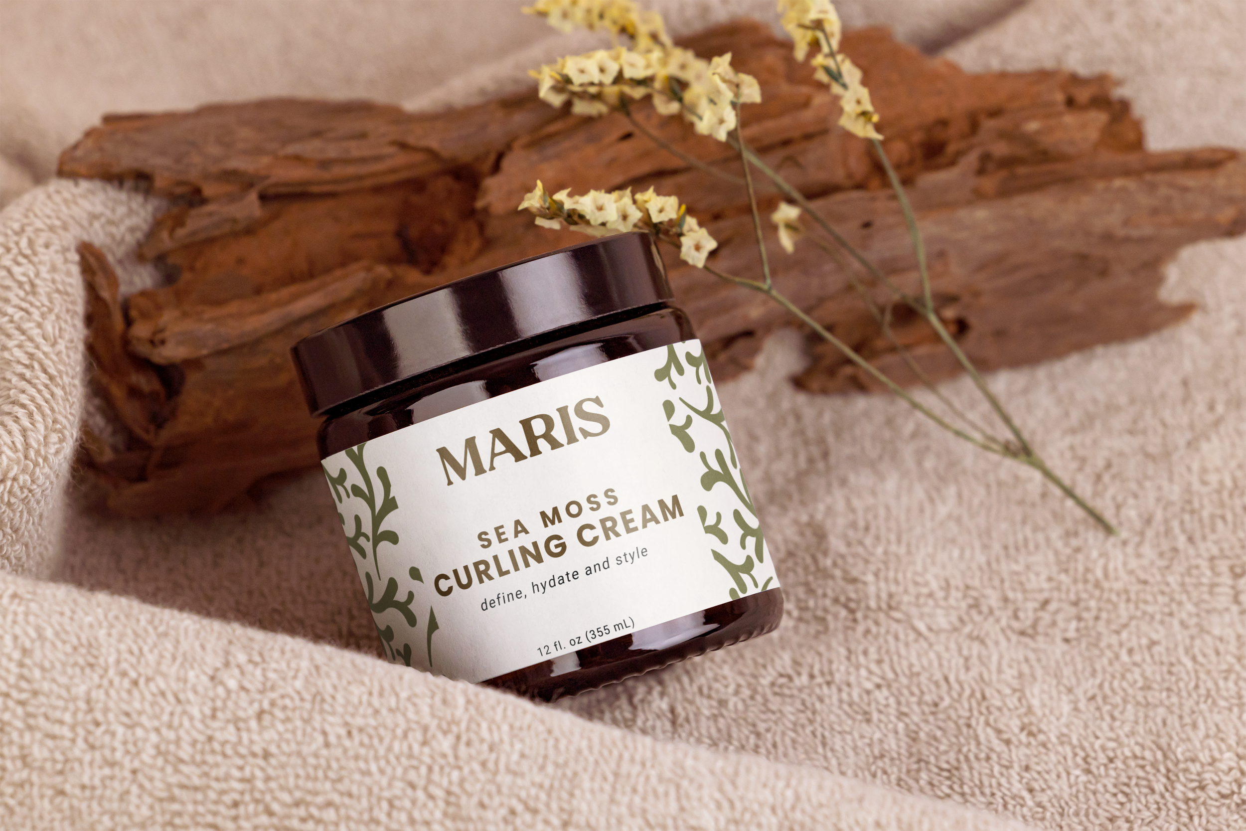 A brown jar of Sea Moss Curling Cream with a white label, placed on a beige textured fabric with a piece of driftwood and a sprig of dried yellow flowers in the background.