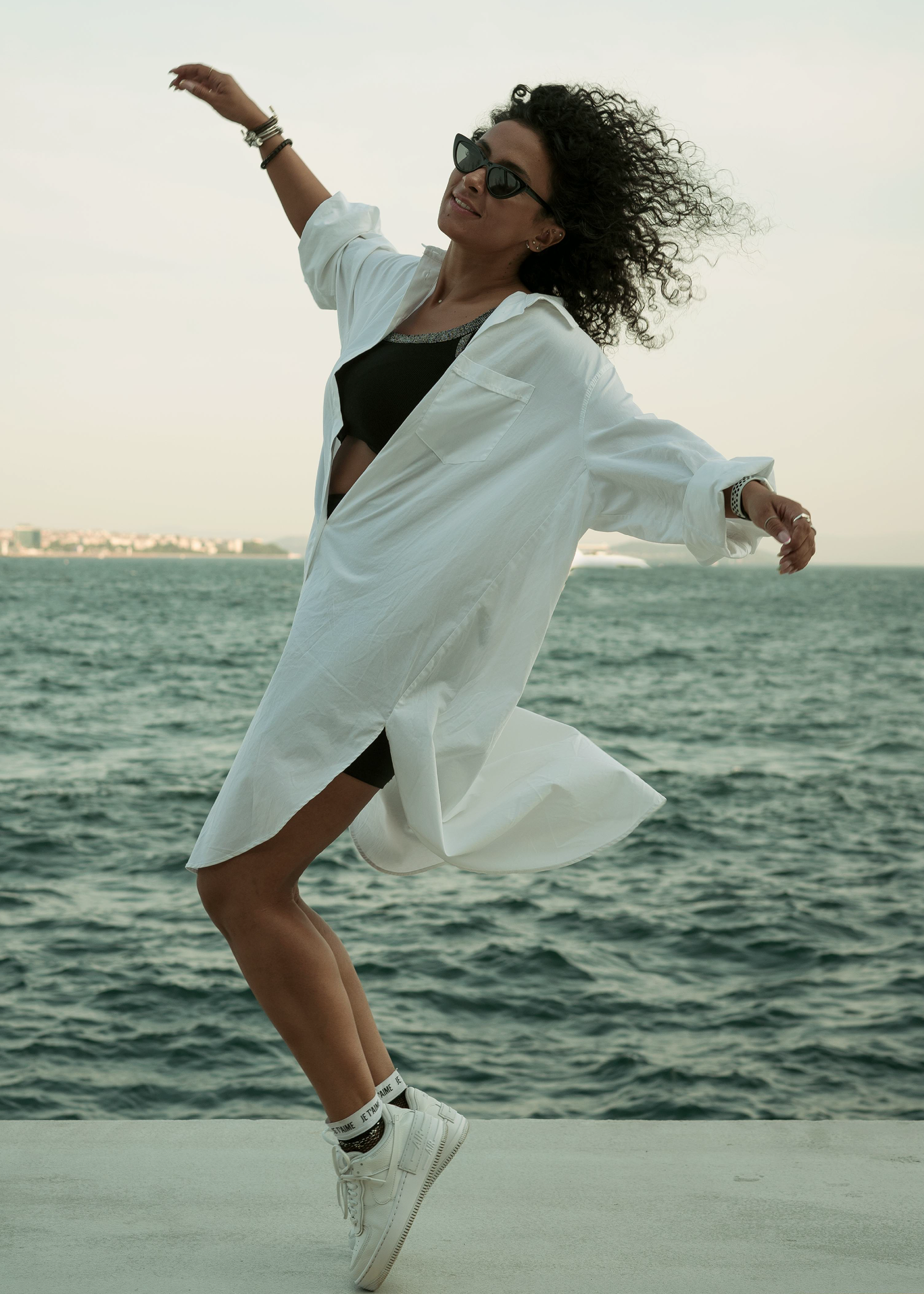 A woman wearing sunglasses, a black crop top, a white button-up shirt, white sneakers, and sock with text is dancing by the water on a gray surface with a cityscape in the background.