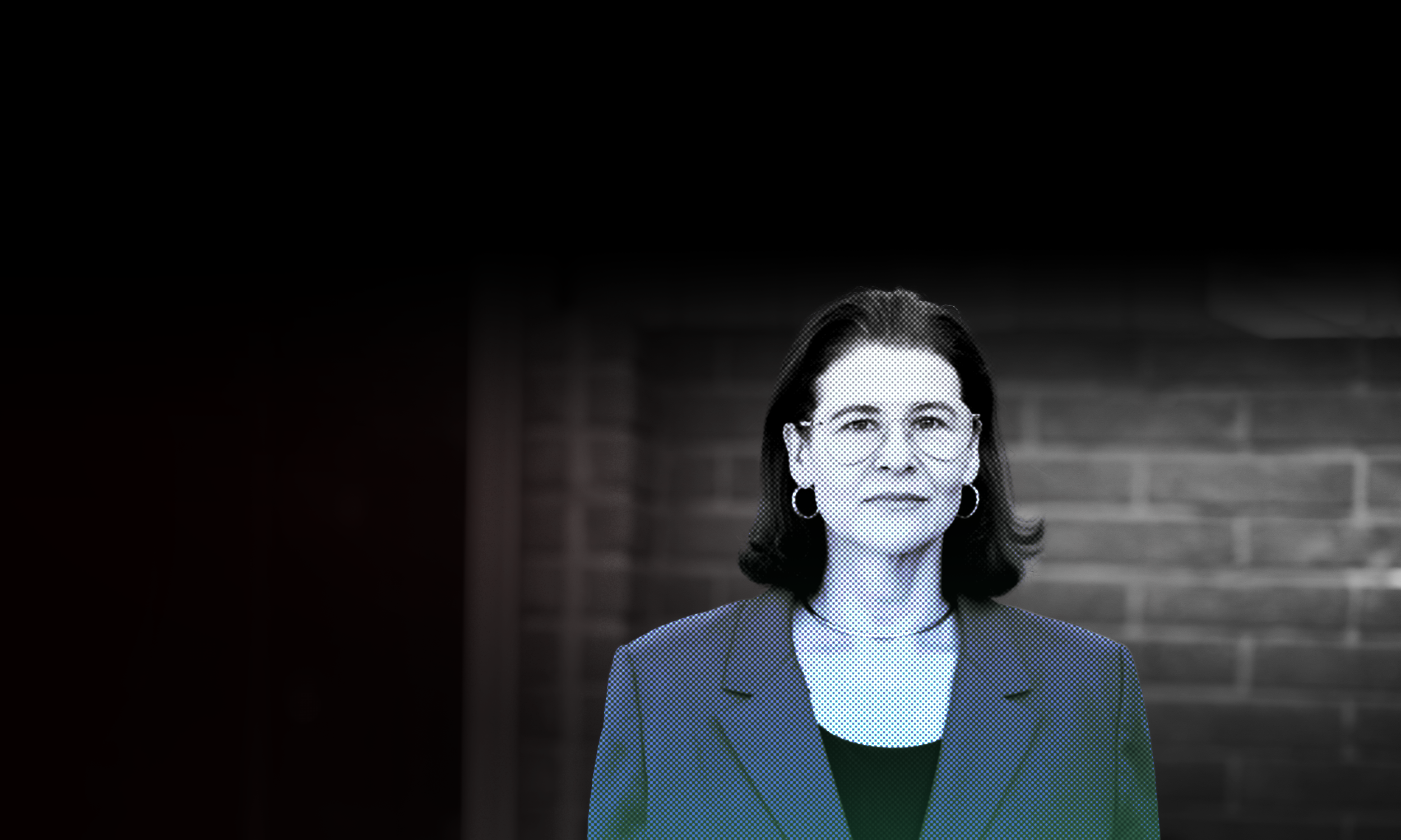 A woman with dark hair, wearing glasses and a dark blazer, standing in front of a brick wall.