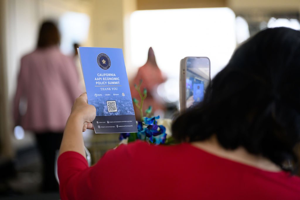 Person holding a blue pamphlet that reads 'California AAPI Economic Policy Summit, Thank You' and is taking a photo of it with a smartphone, at an indoor event.