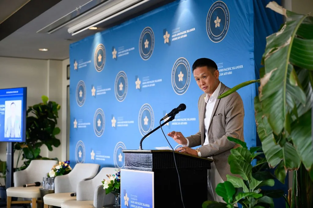 Man in a beige blazer speaking at a podium during an event at the Asian and Pacific Islander American Affairs conference, with a blue backdrop and potted plants nearby.
