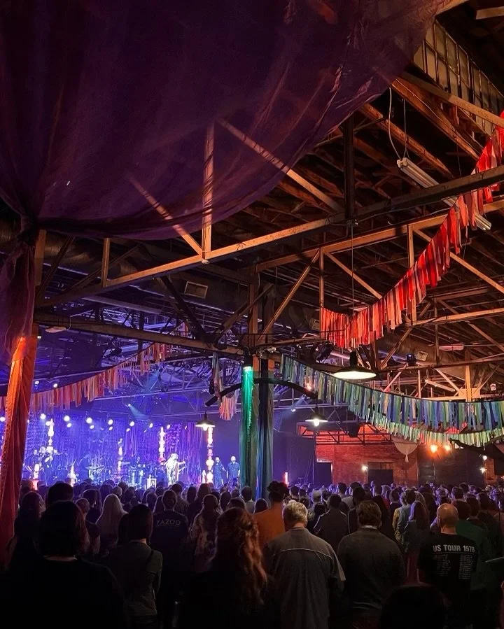Indoor concert with a large crowd facing a stage with musicians and performers, colorful hanging decorations, and stage lighting.
