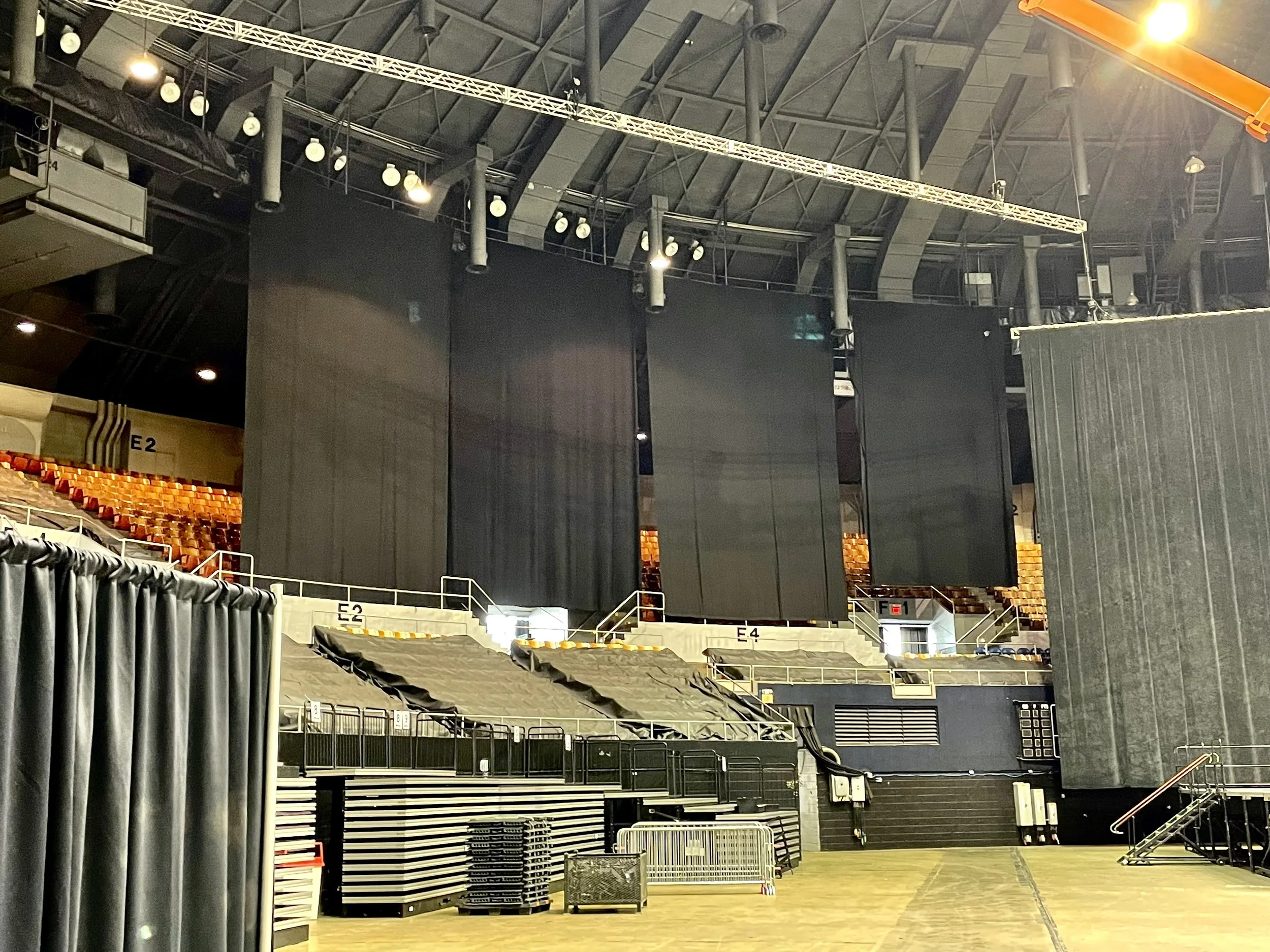 An indoor sports arena with empty seating, scaffolding, and large black drapes hanging from the ceiling, likely set up for an event or concert.