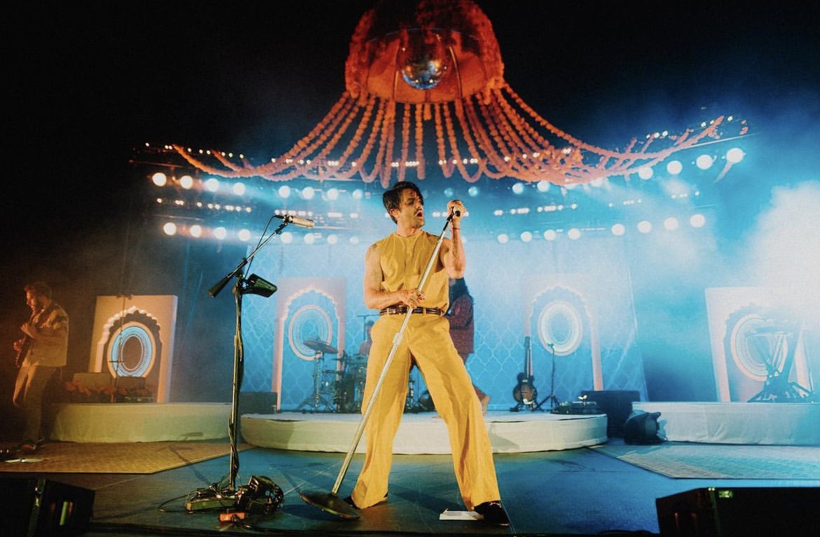 Performer singing on stage in a yellow outfit, holding a microphone stand, with colorful stage lighting, large custom fabric backdrop, and musical instruments in the background.