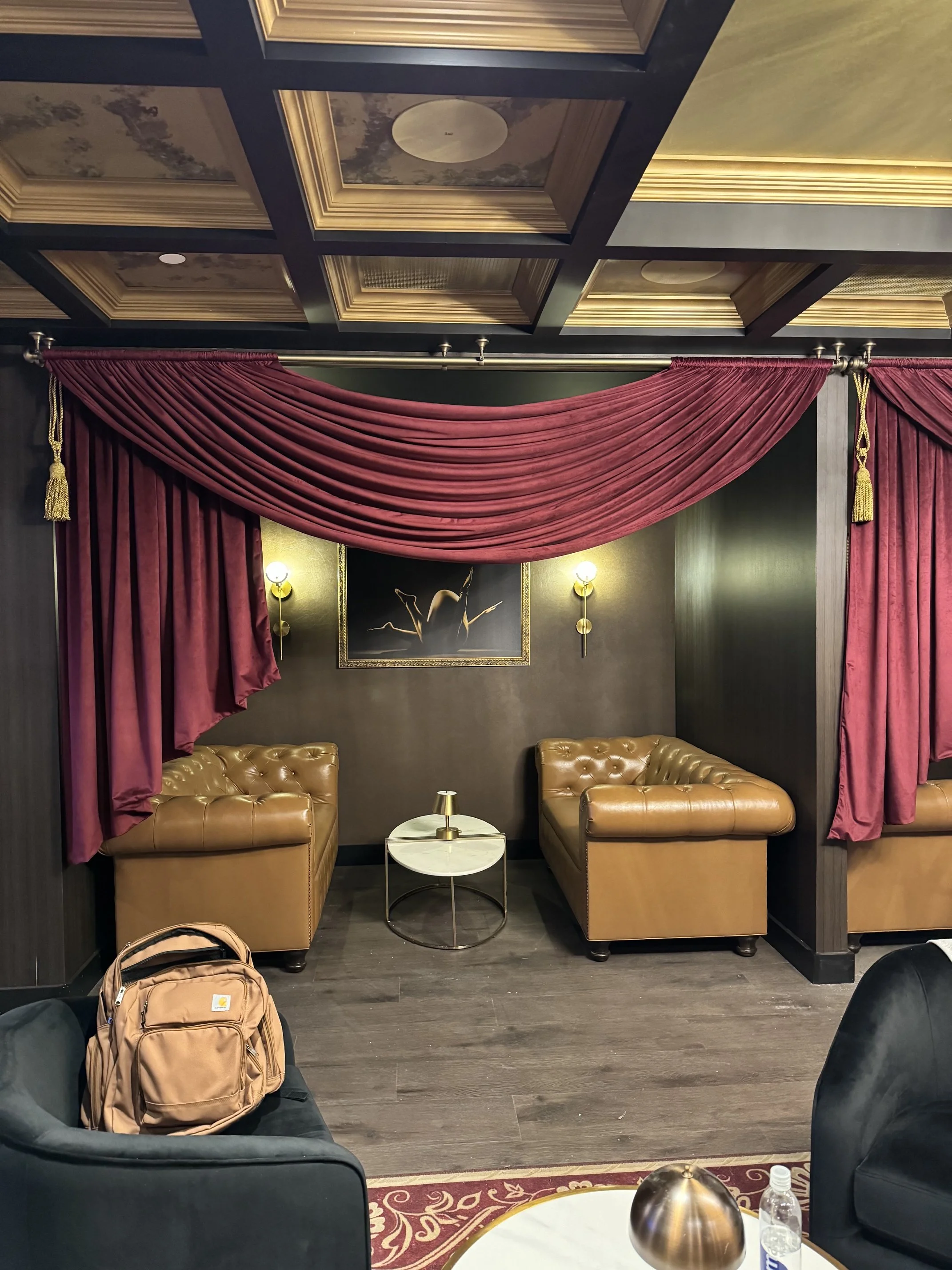 A cozy lounge with dark walls and elegant red curtains framing a view of two tan leather armchairs separated by a round marble-topped side table. Overhead, a detailed painted ceiling and a framed picture of ballet dancers add artistic touch. A backpack rests on a black armchair in the foreground, and a small table with a metallic lamp and water bottle is visible.