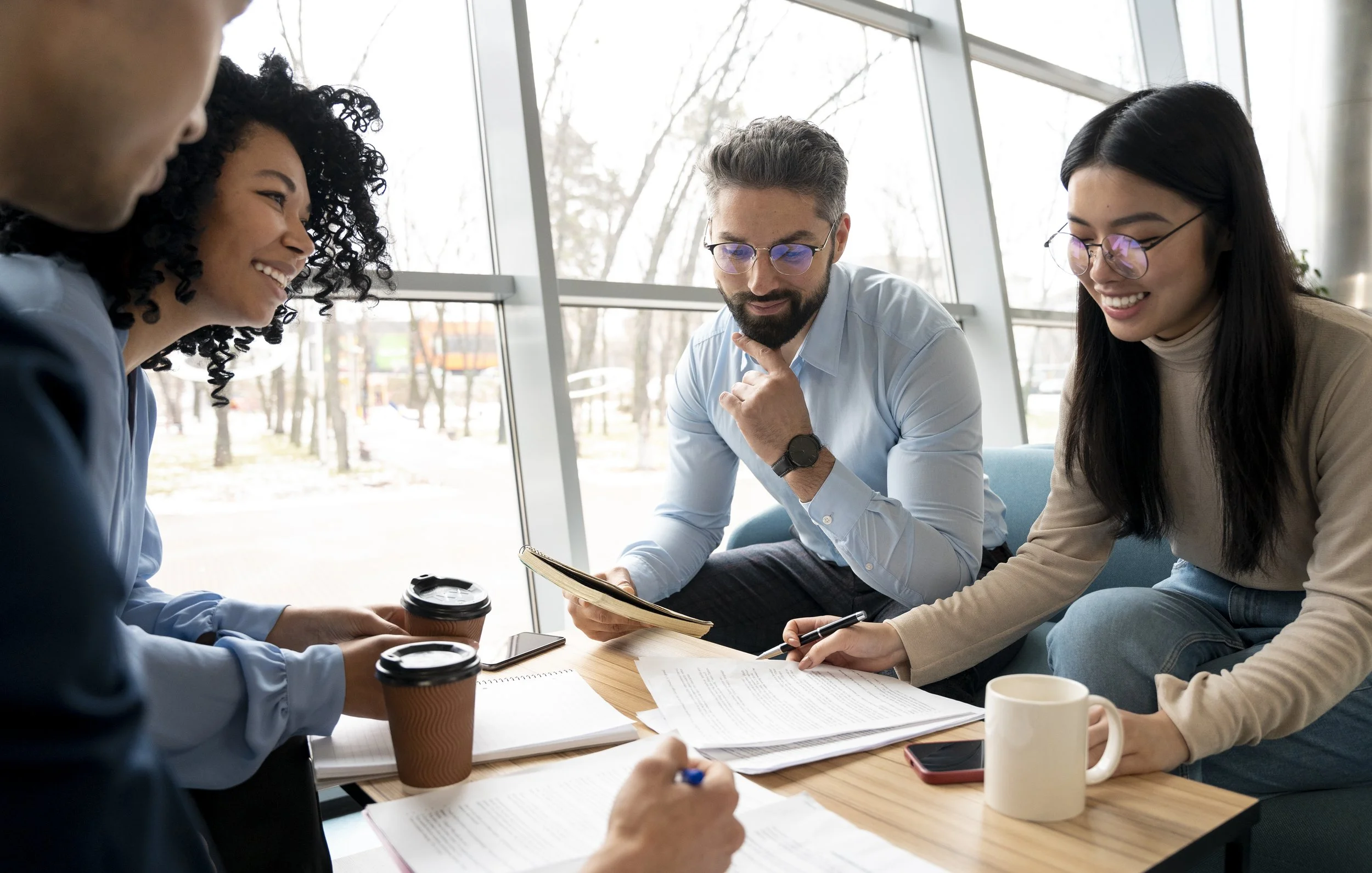Group of four diverse people sitting around a table in a bright room, engaged in discussion and looking at documents and a tablet. They have coffee cups on the table.