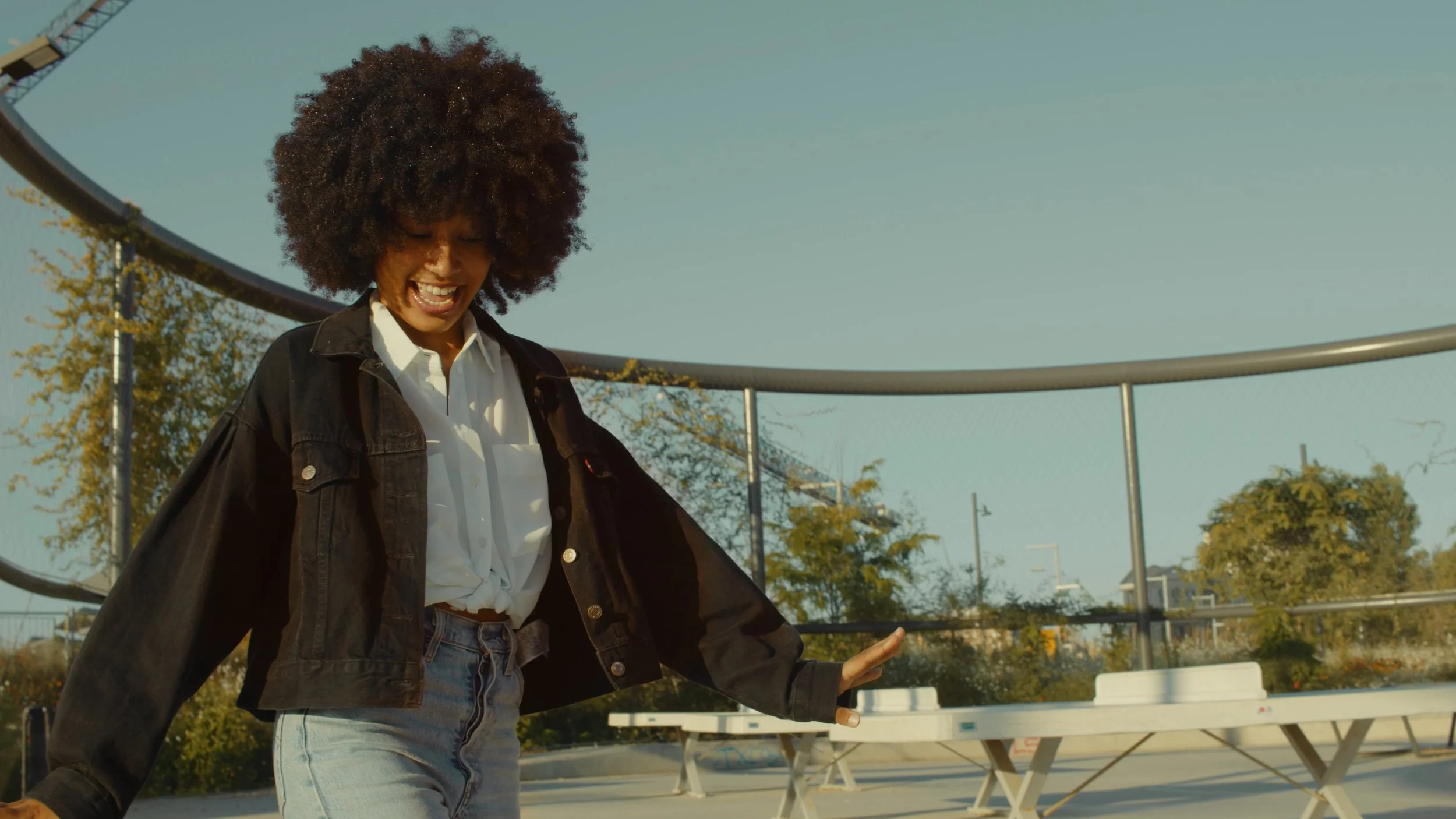 A woman with curly hair laughing and enjoying in an outdoor skate park during daytime, with skate ramps and trees in the background.