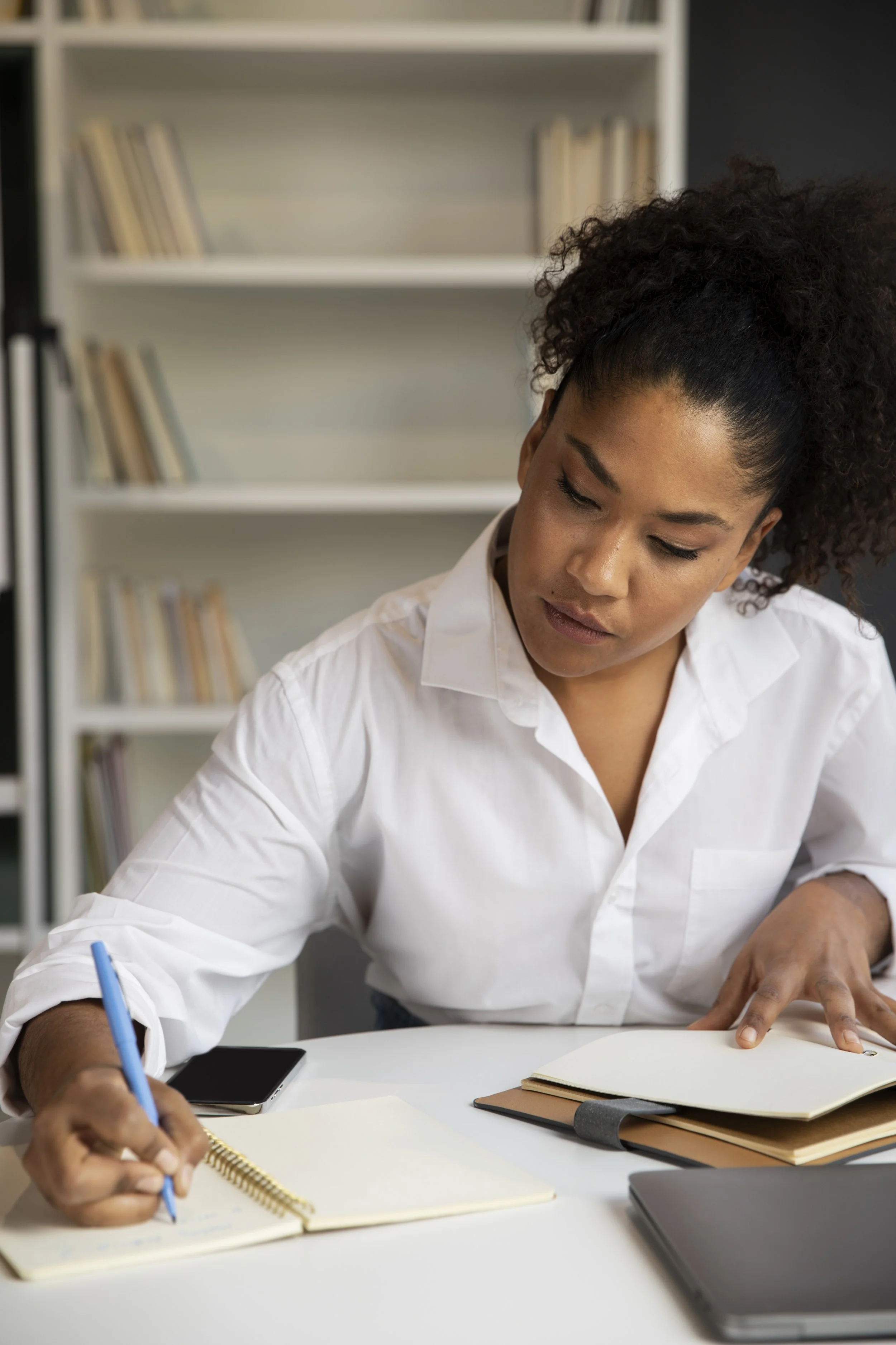 A woman with curly hair tied up in a ponytail sitting at a desk, writing in a notebook with a blue pen. She is wearing a white shirt, and there are multiple notebooks, a laptop, a smartphone, and a tablet on the desk. A white bookshelf with books is in the background.